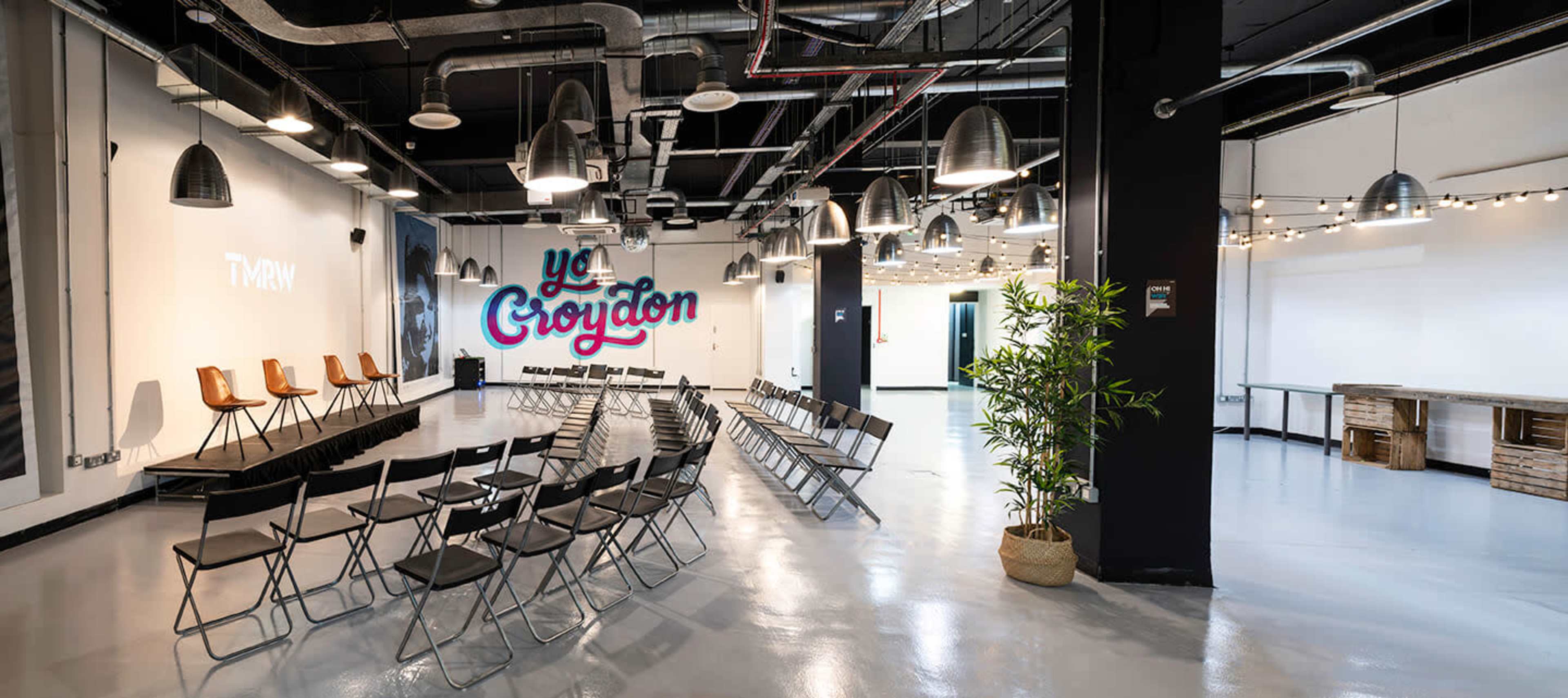 An indoor venue set up with rows of folding chairs facing a wall that has the words "Ye Croydon" painted on it, along with a few tables and a potted plant in the corner.
