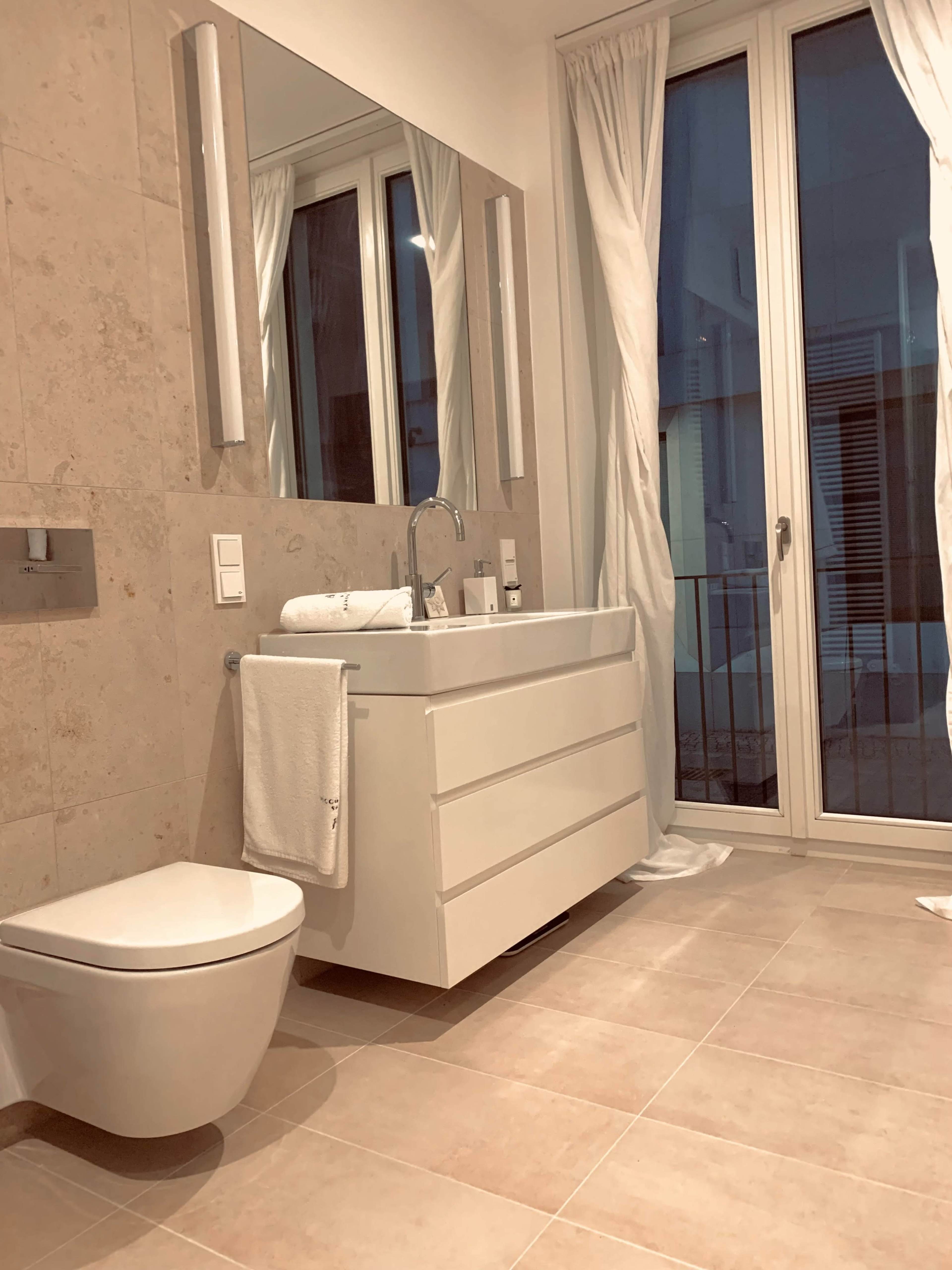 A modern bathroom featuring a white vanity with a large mirror, a toilet, and floor-to-ceiling windows covered by sheer curtains.