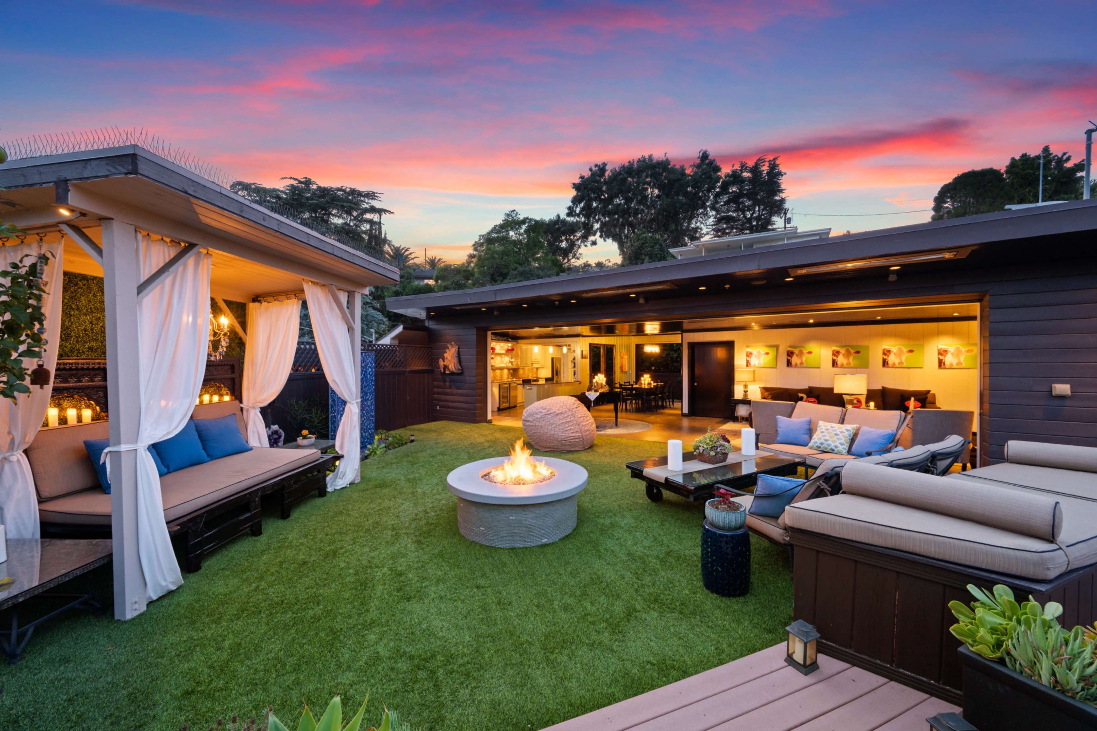 A backyard patio features a central fire pit surrounded by seating, with two covered lounge areas and a backdrop of a colorful sunset sky.