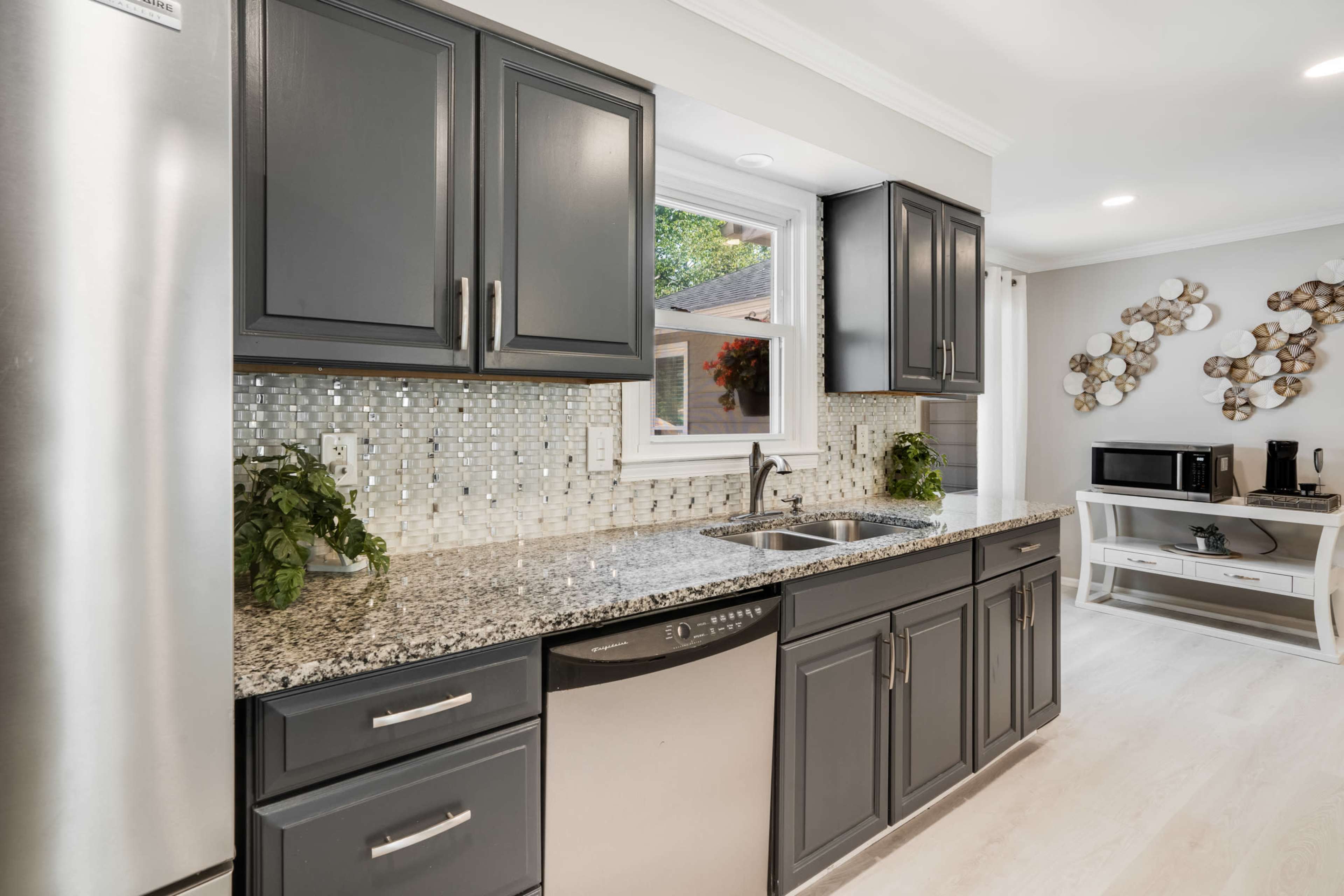 A modern kitchen with dark cabinetry, a stainless steel refrigerator, a granite countertop, and a window overlooking a garden.