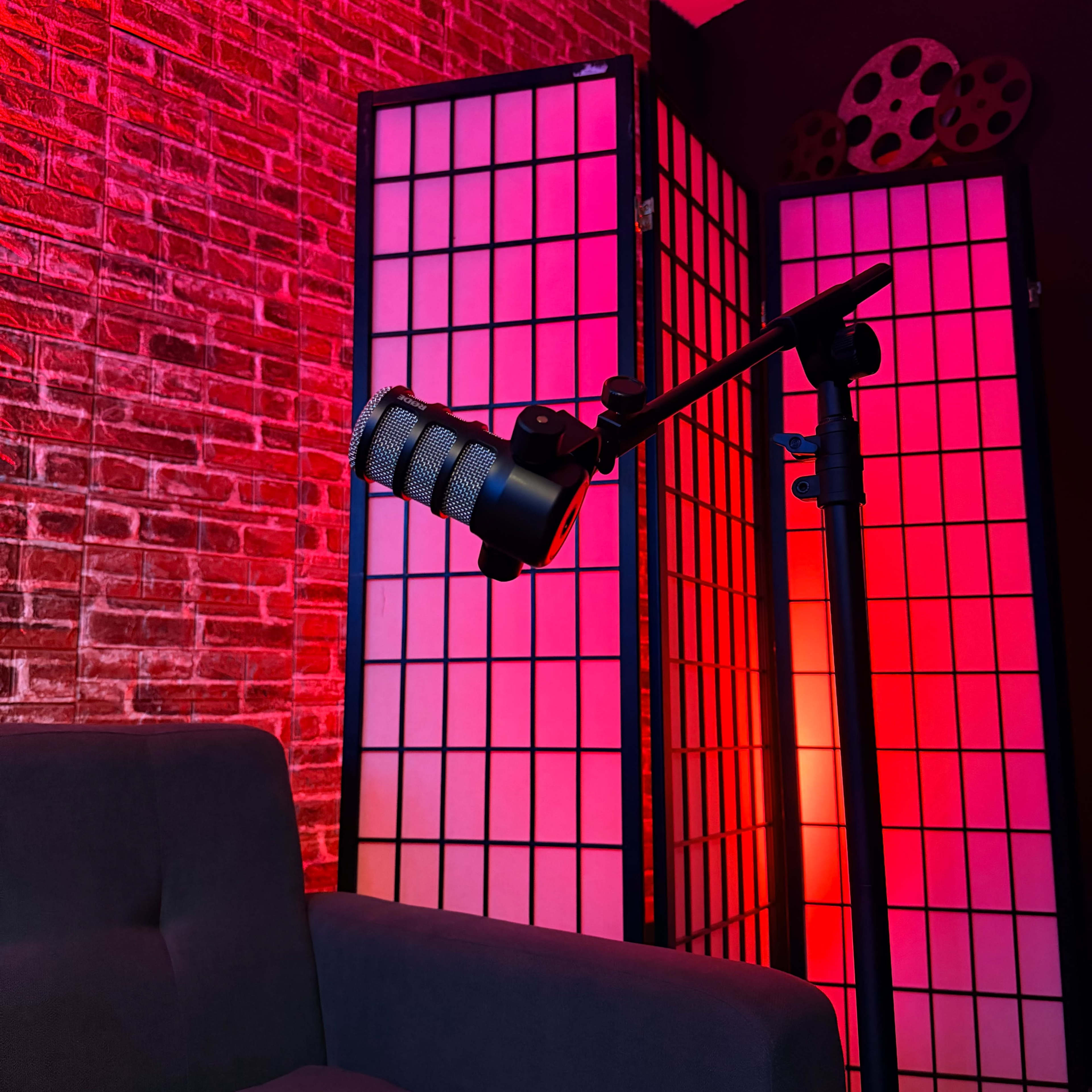 A black microphone is mounted on a stand in front of a red and pink-lit backdrop created by a shoji screen and brick wall.