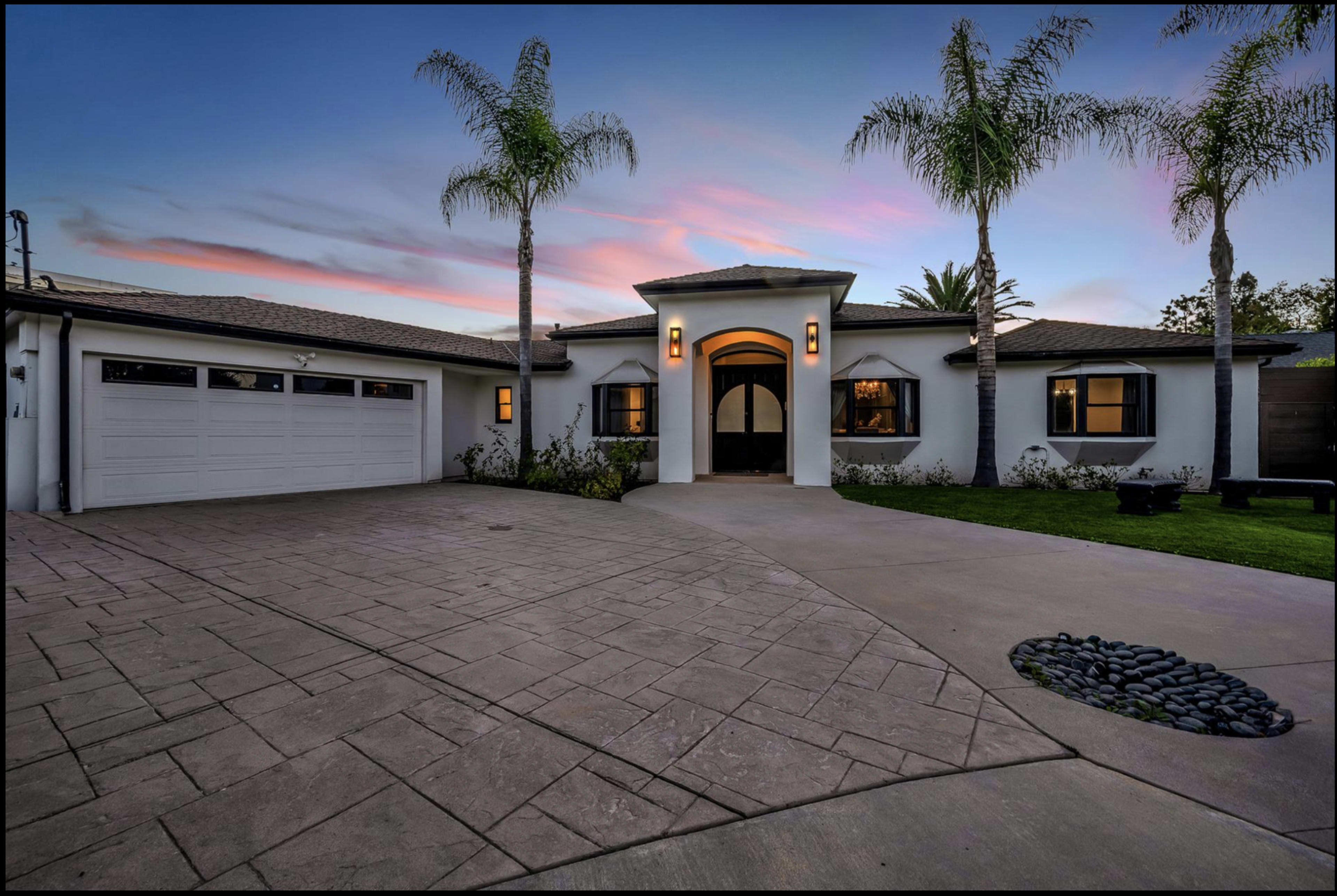 A modern single-story house with a circular entryway, palm trees, and a patterned driveway under a colorful sky at dusk.