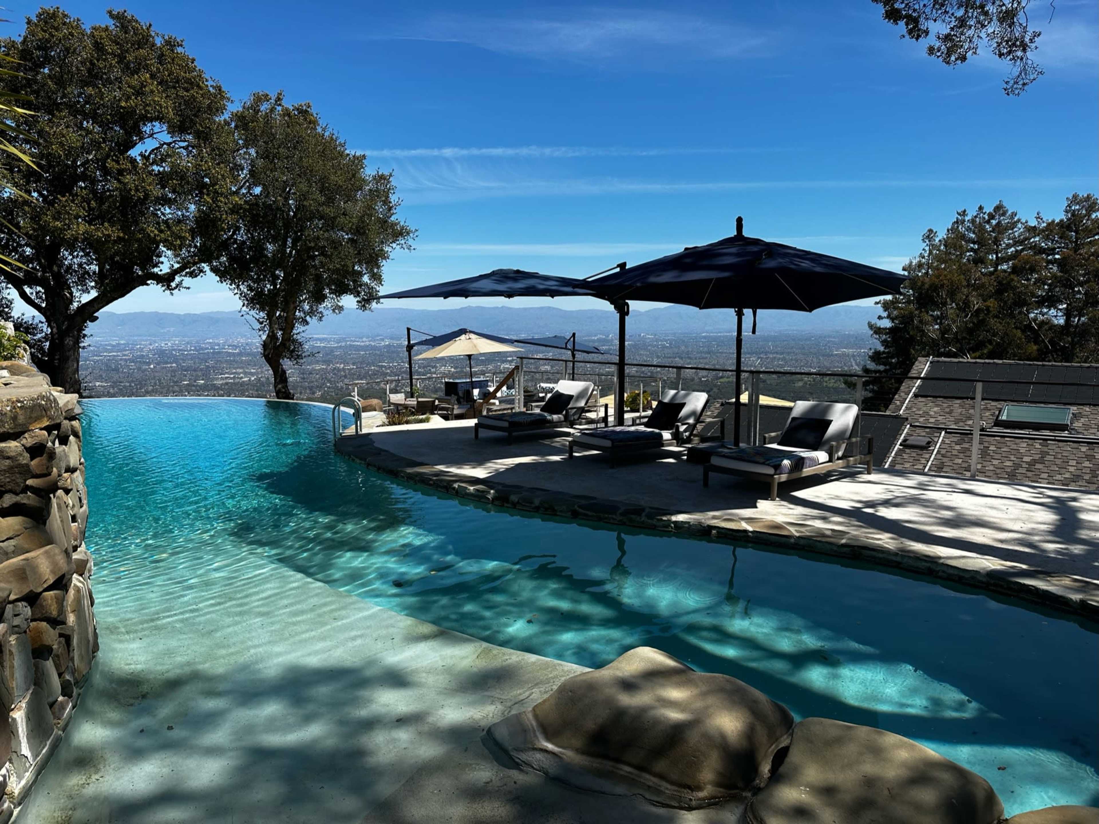 The image shows a curved infinity pool overlooking a valley, with sun loungers and umbrellas beside it under a clear blue sky.