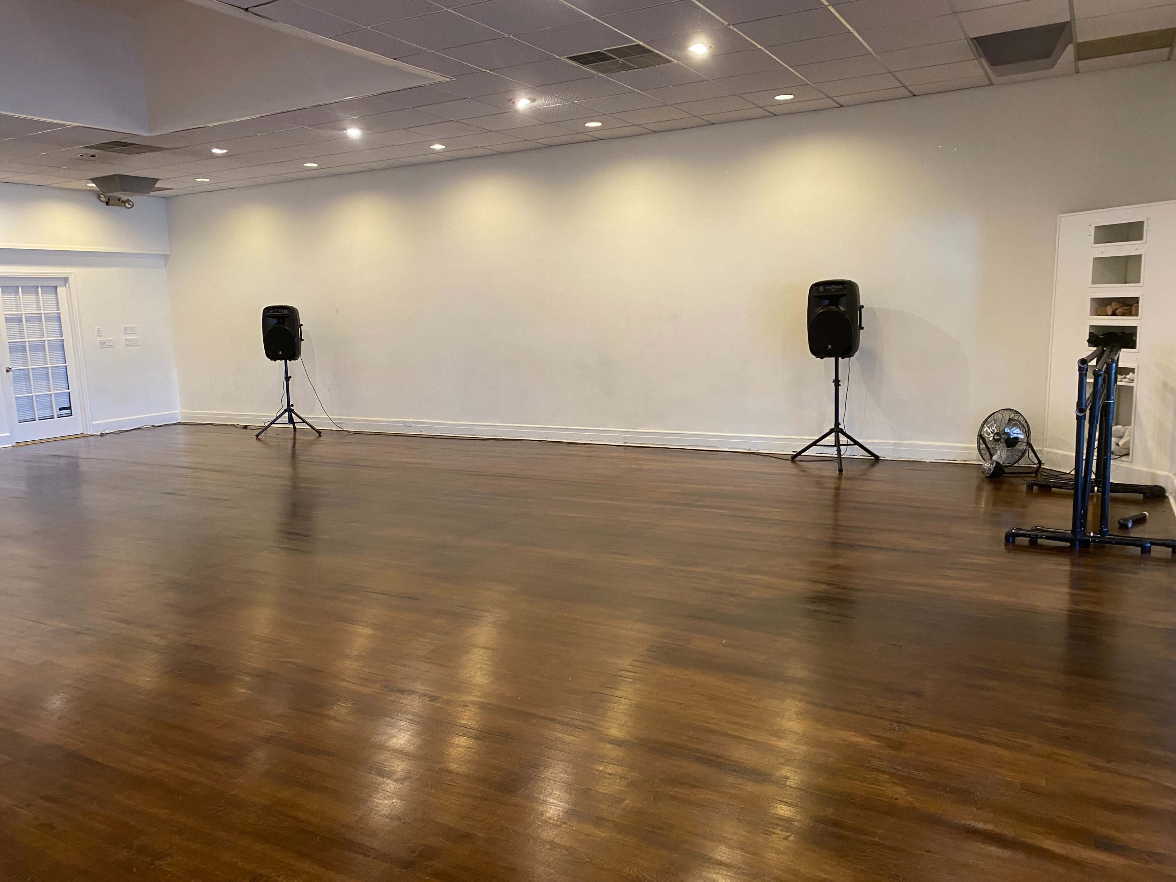 The image shows an empty indoor space with polished wooden floors, two speakers set on stands, and a blue exercise machine against the wall.