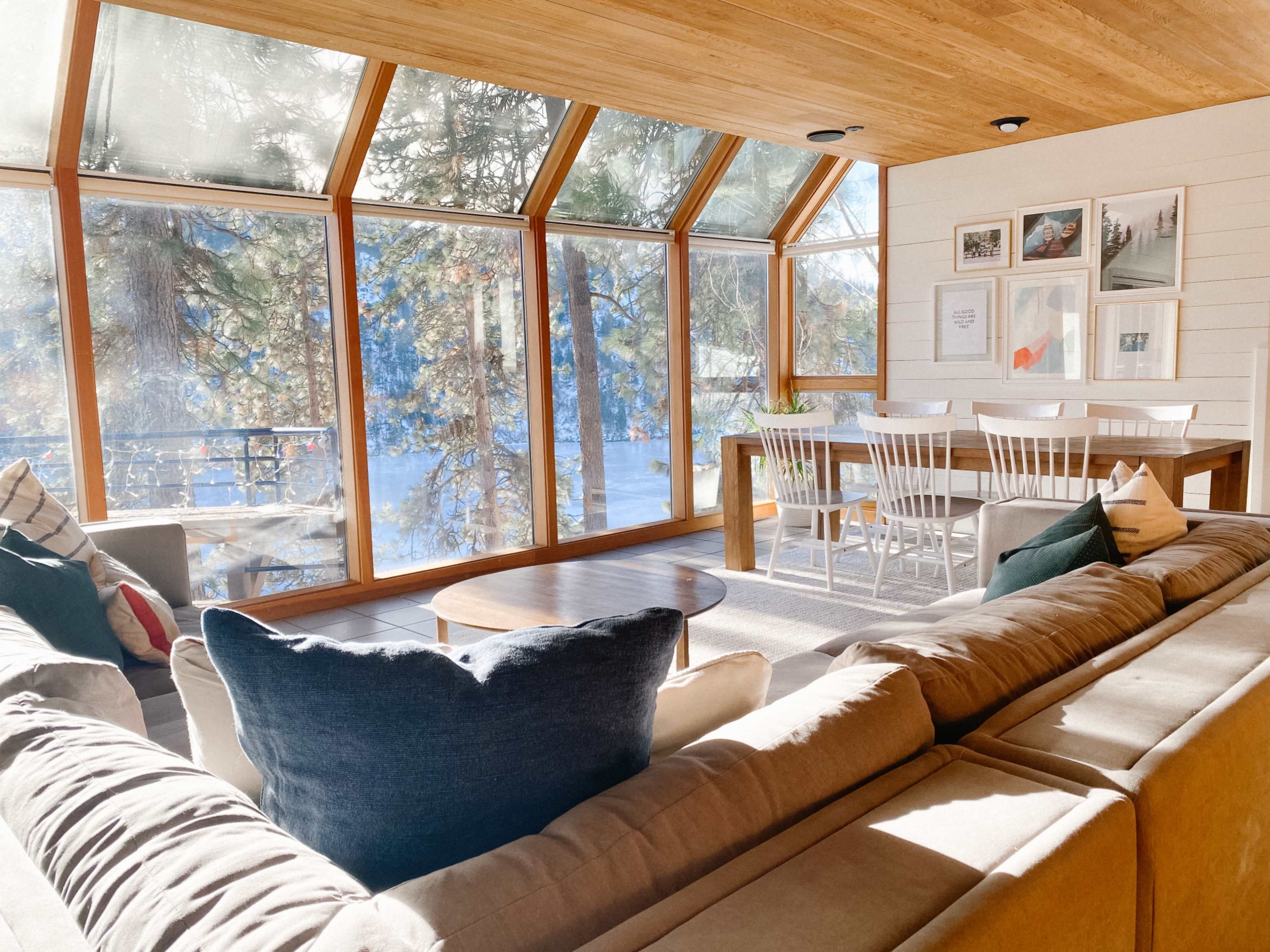 A spacious living room features large windows with a view of trees and a dining area with a table and white chairs.