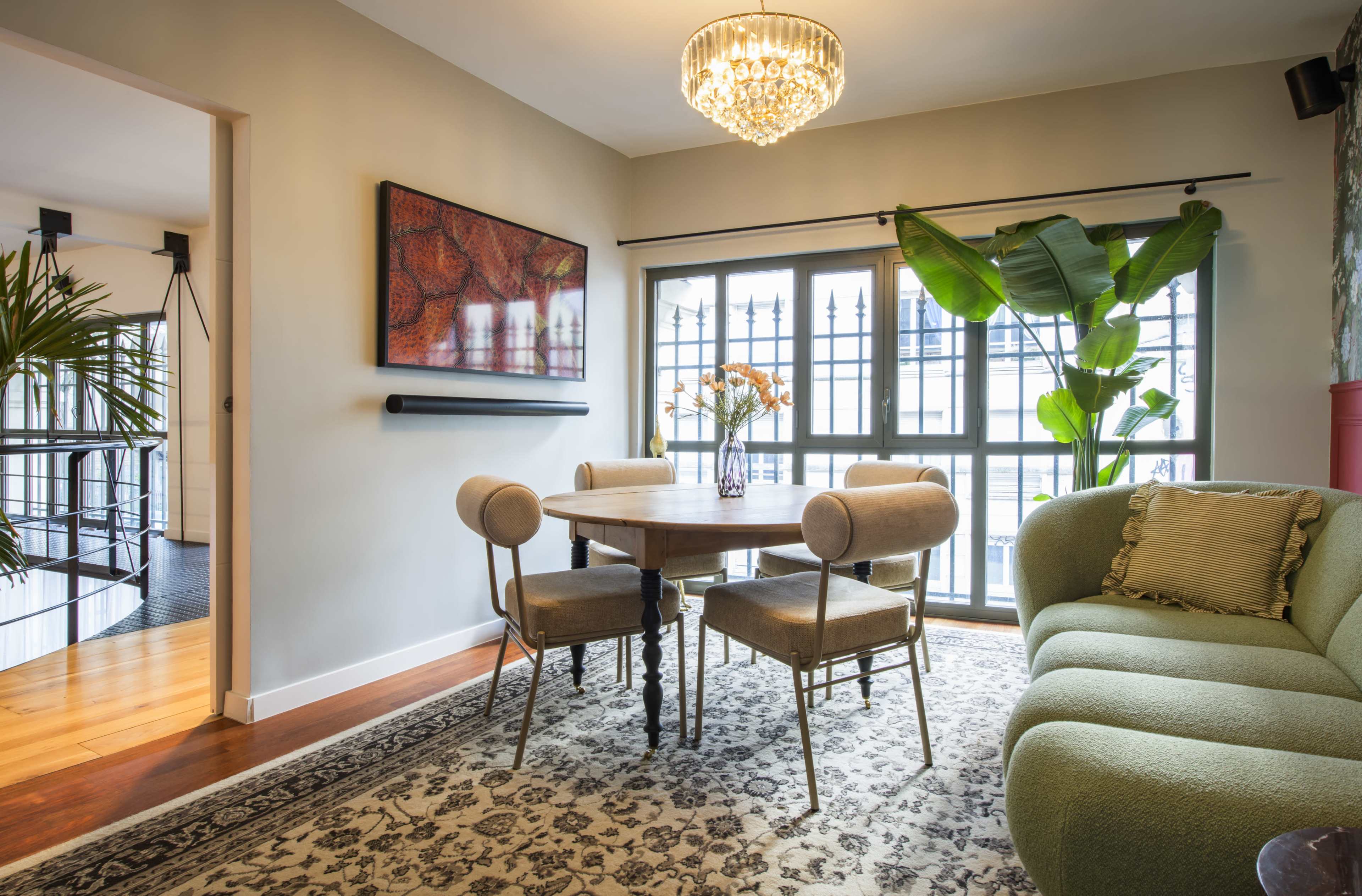 A well-lit dining area features a round wooden table surrounded by tufted chairs, a large potted plant, and a modern chandelier.