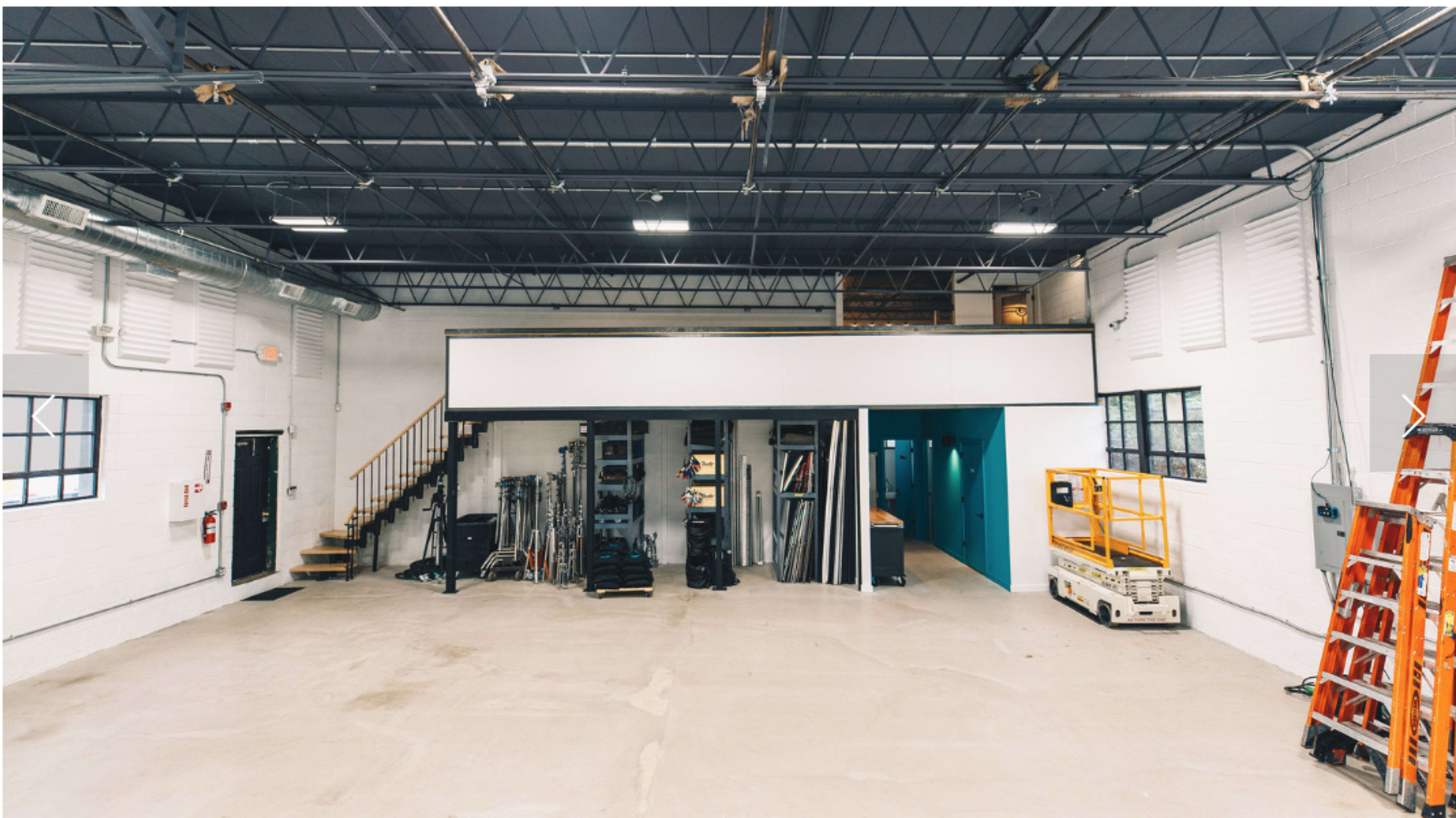 The image shows a spacious industrial workshop with a mezzanine, various equipment, and a ladder against the wall.