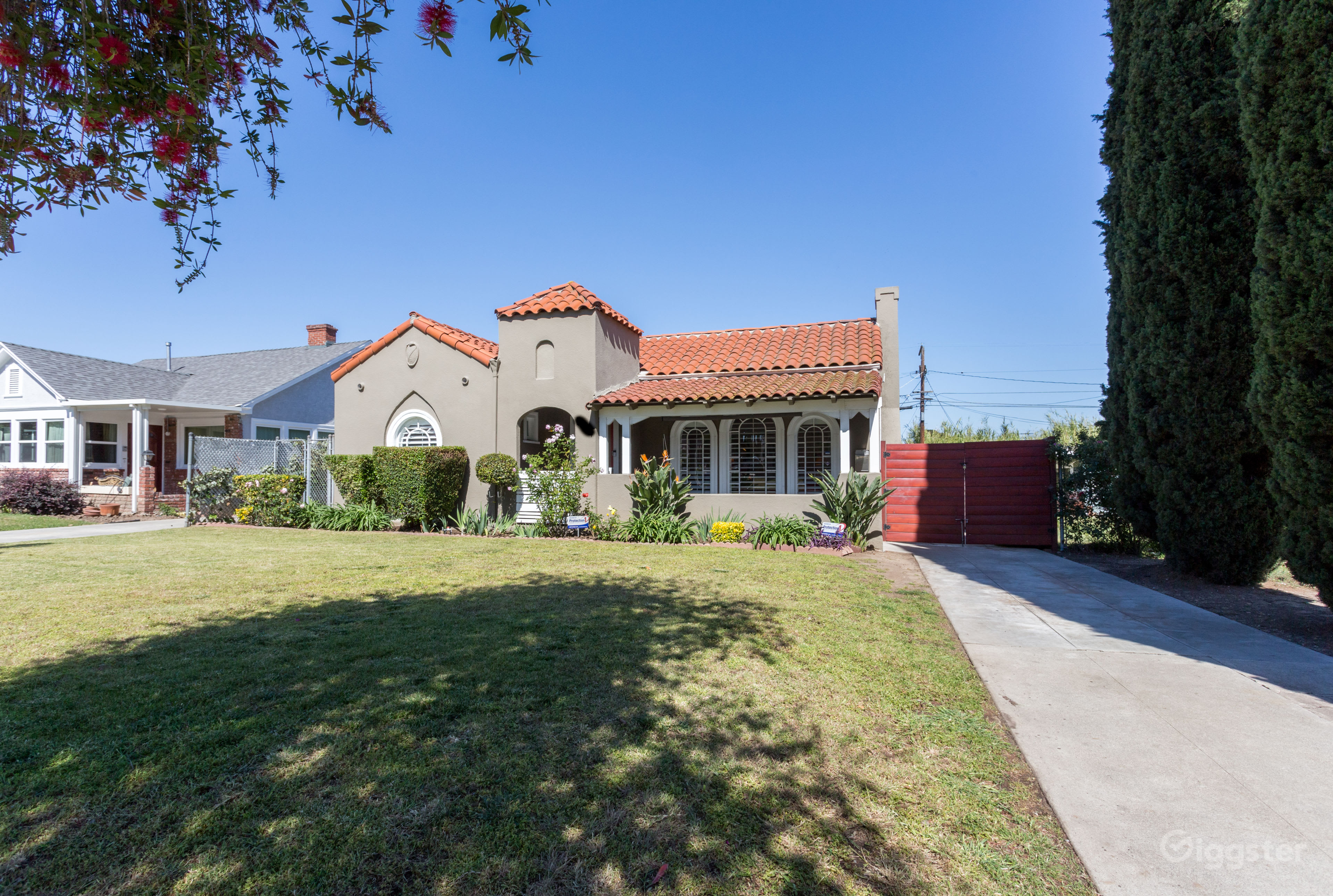 A single-story house with a red tile roof, decorative archways, and a landscaped front yard is situated beside a concrete driveway.