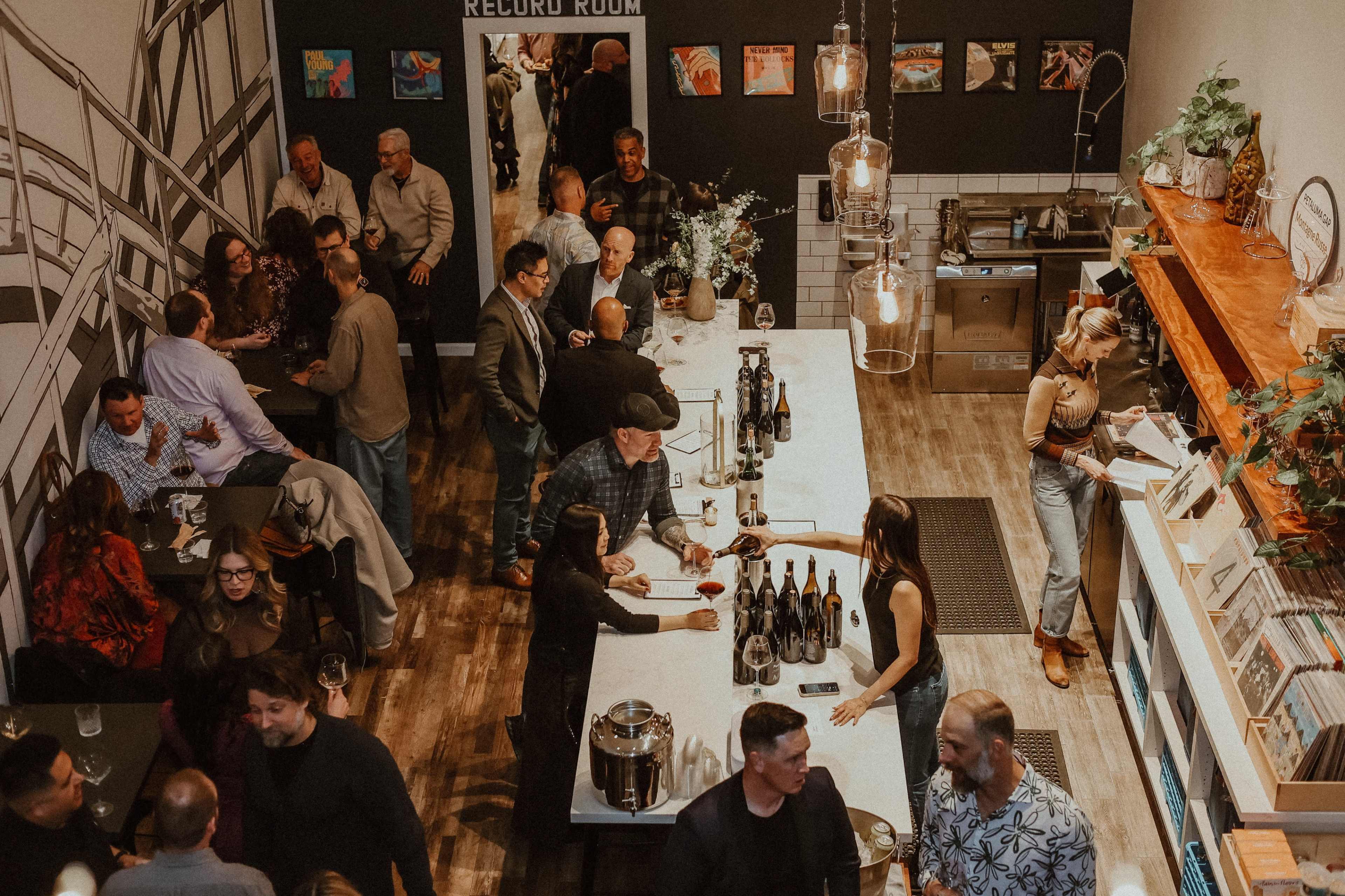 A lively gathering takes place in a bar, with patrons socializing around a long counter and various artworks displayed on the walls.