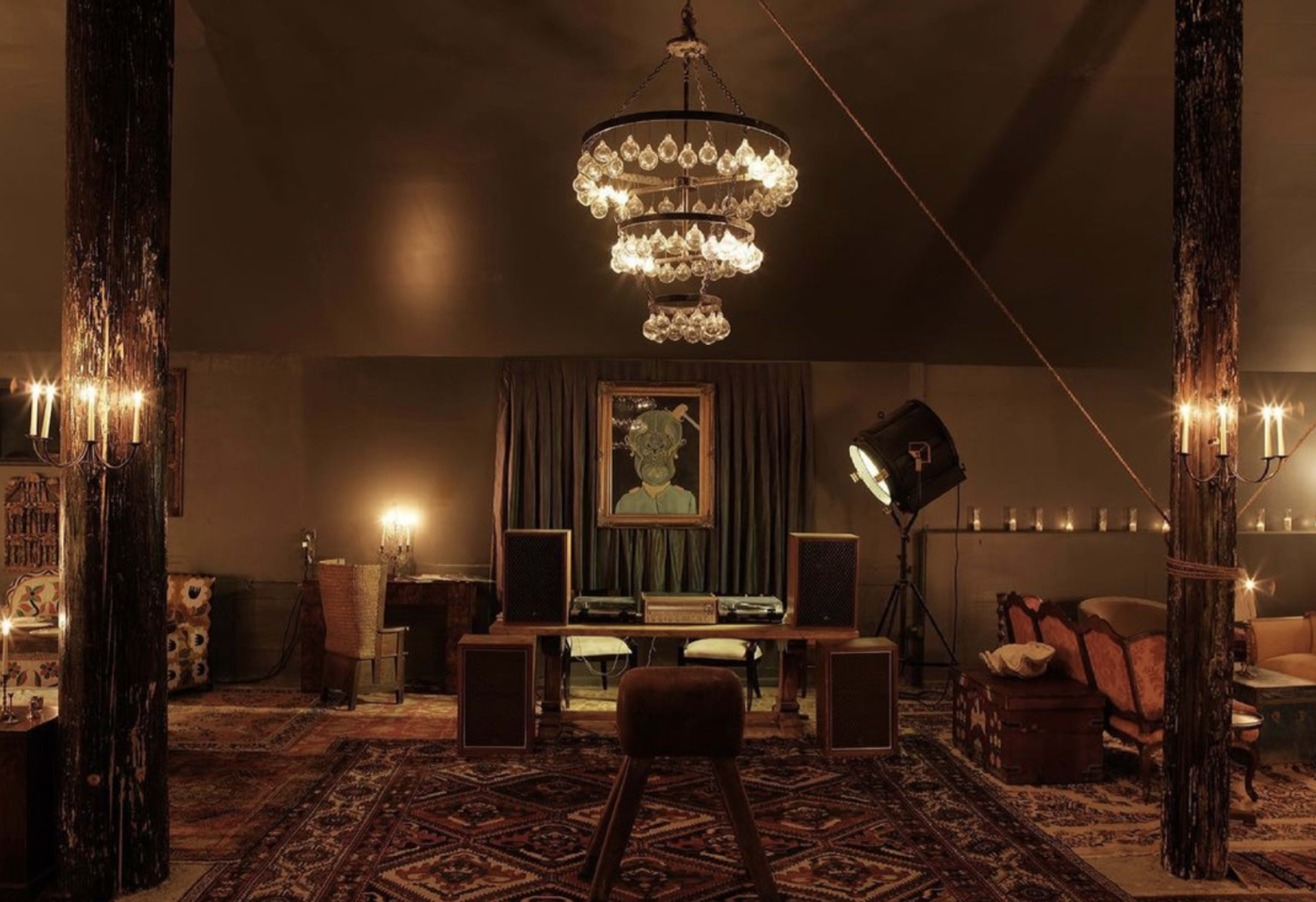 The scene features a dimly lit room with a large chandelier, wooden beams, a decorative rug, and a vintage seating area arranged around a central table with audio equipment.