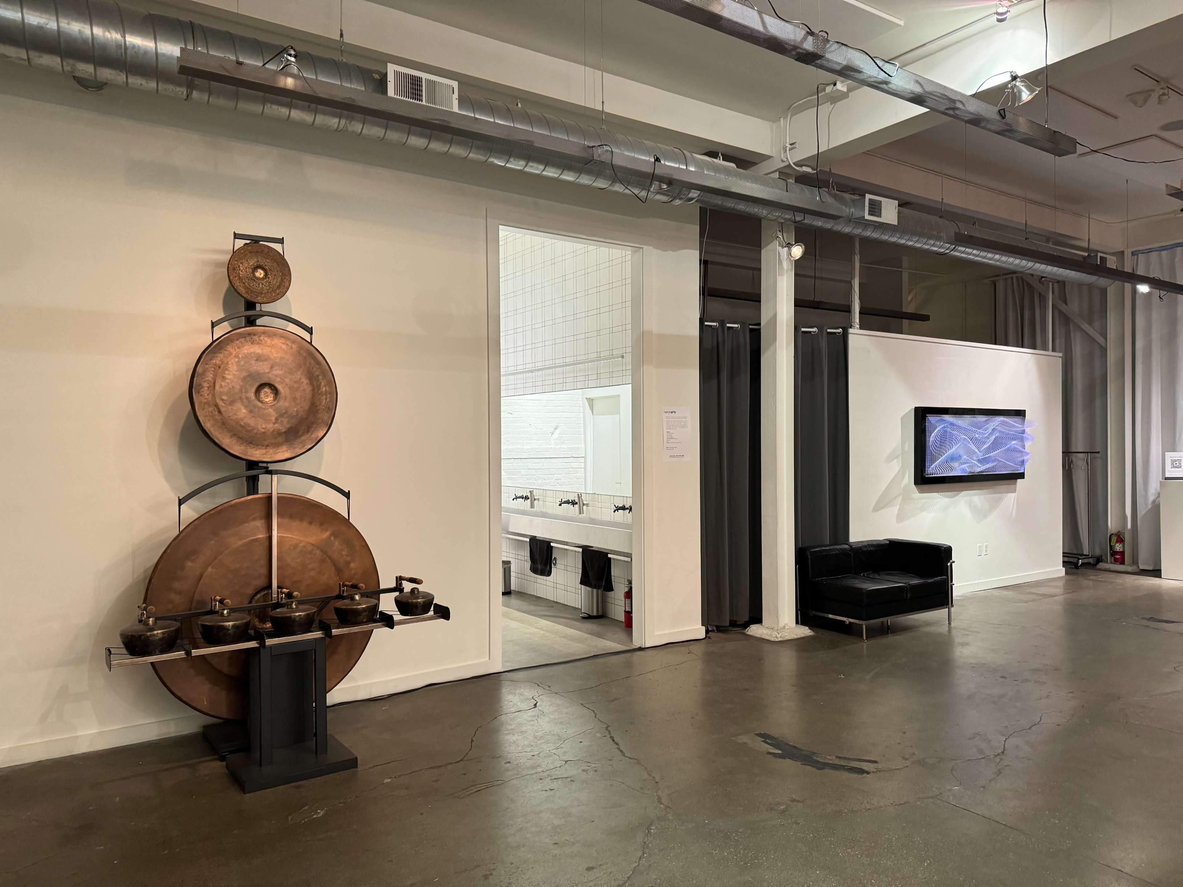 The image shows a contemporary art gallery featuring a large circular sculpture made of metal on the left and a dark seating area with a wall-mounted screen on the right, with a bathroom entrance visible in the center.