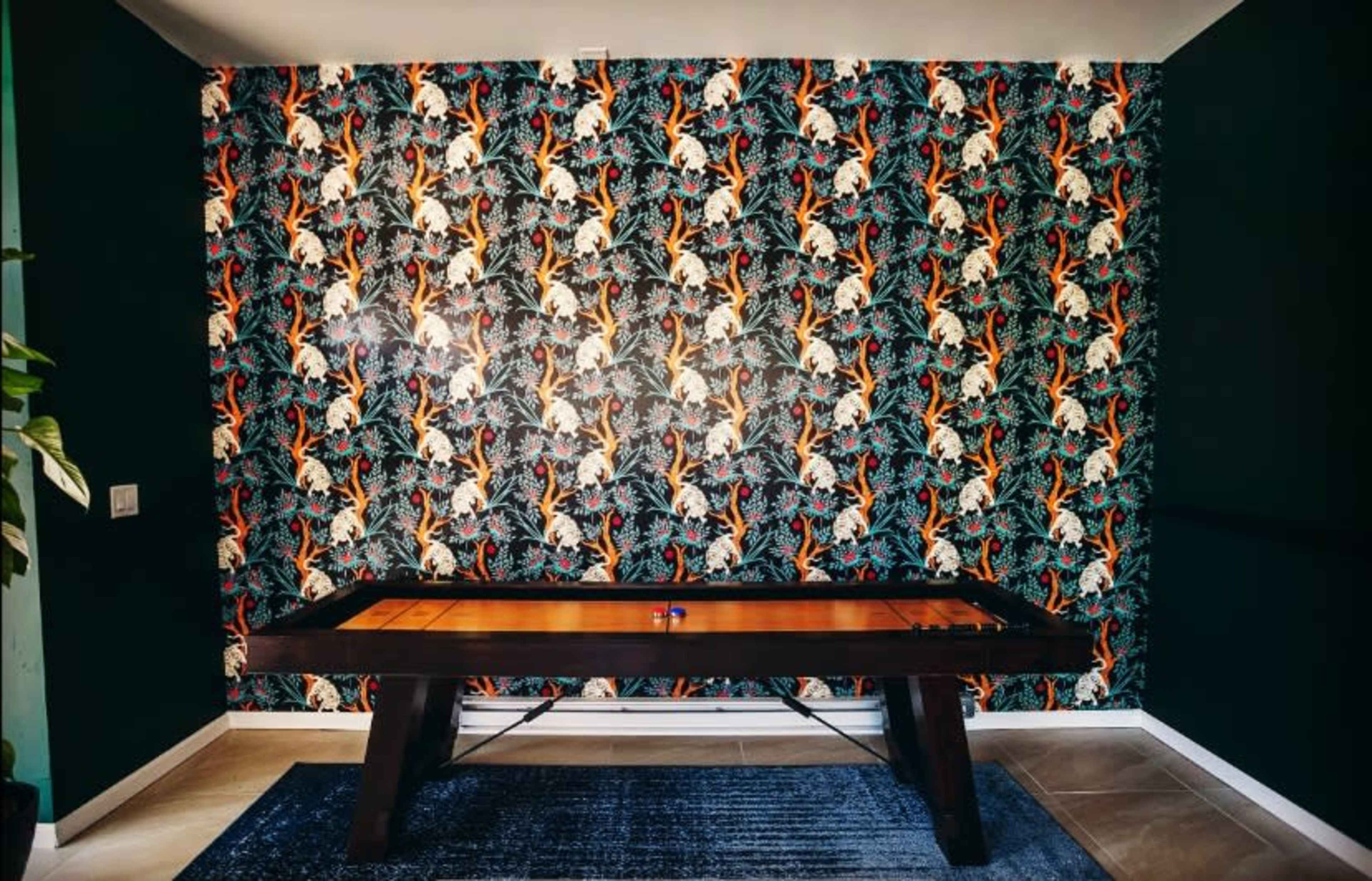 A pool table is set against a vibrant, patterned wallpaper decorated with tropical motifs, surrounded by dark green walls.