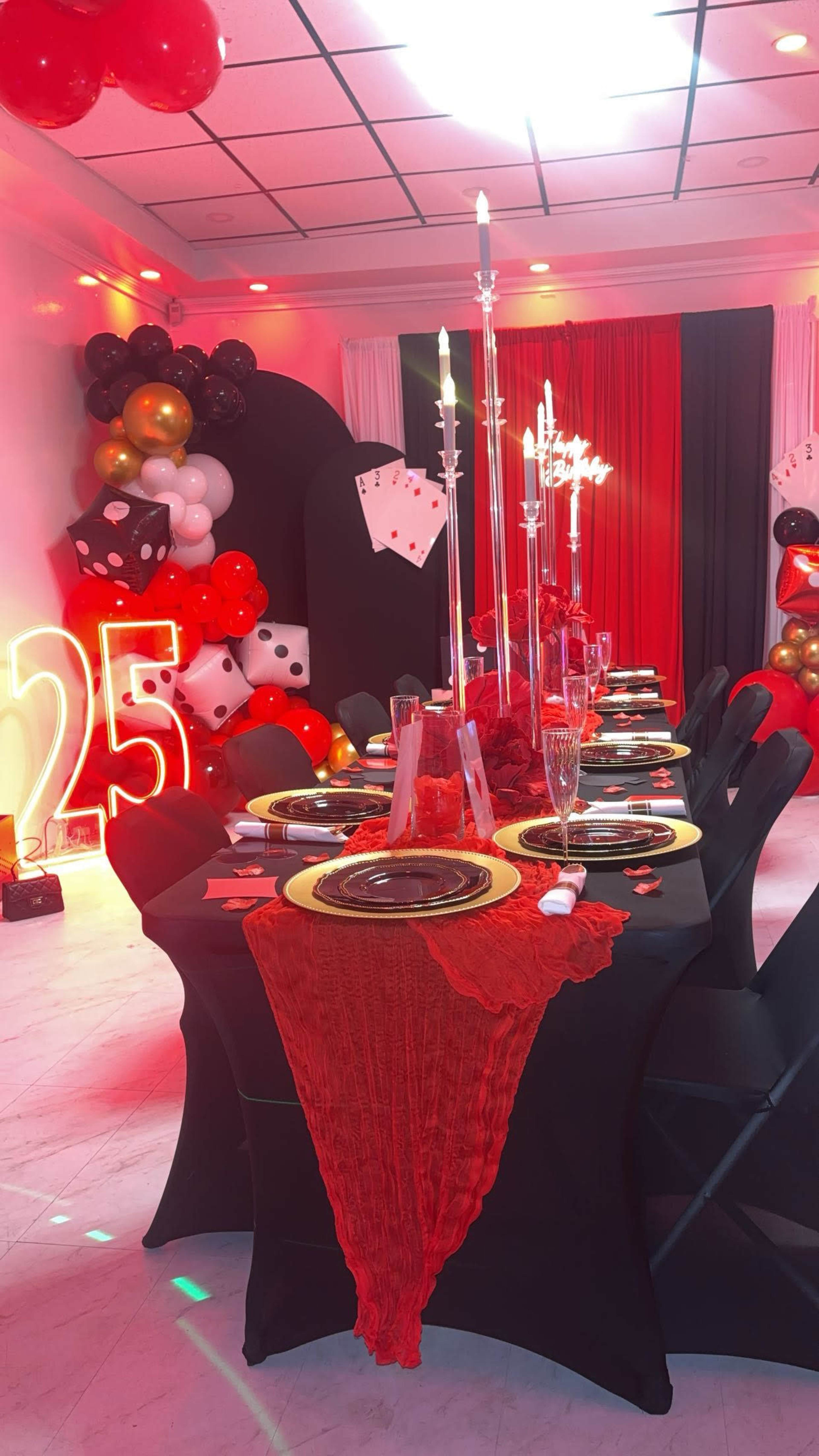 A banquet table is elegantly set with black cloths, gold plates, and candle holders, surrounded by red and black decorations, symbolizing a 25th celebration.