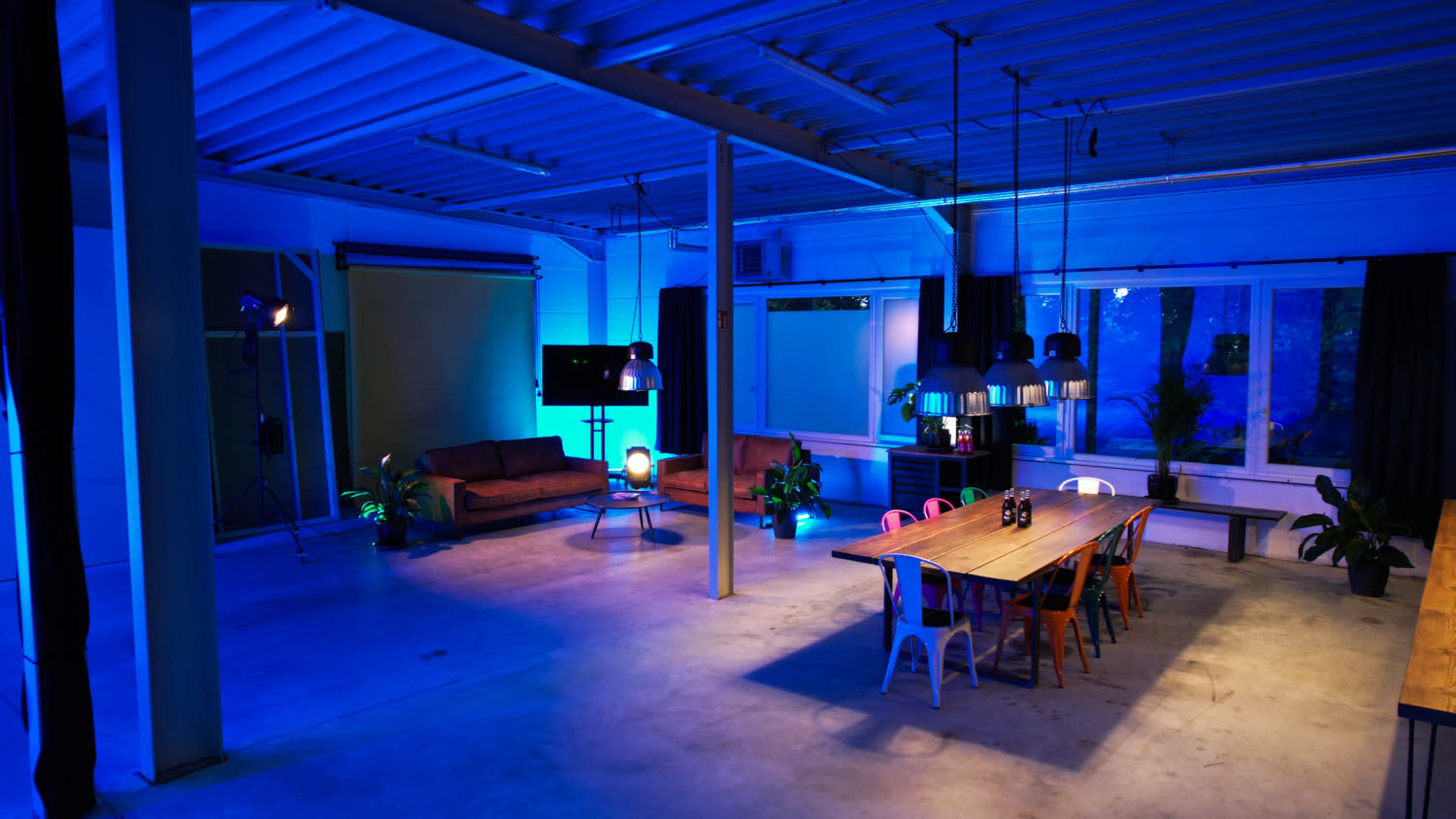 A spacious, modern room with a long wooden table, colorful chairs, a couch arrangement, and blue lighting illuminating the space.