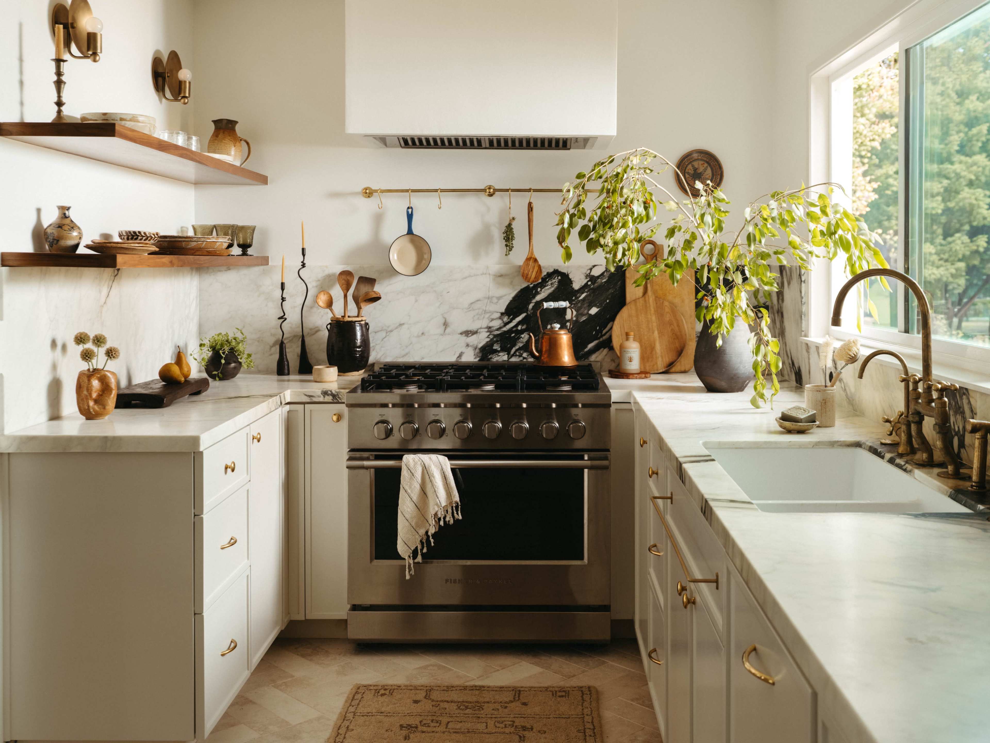 The image shows a modern kitchen featuring a marble countertop, stainless steel stove, and open shelves with decorative items and plants.