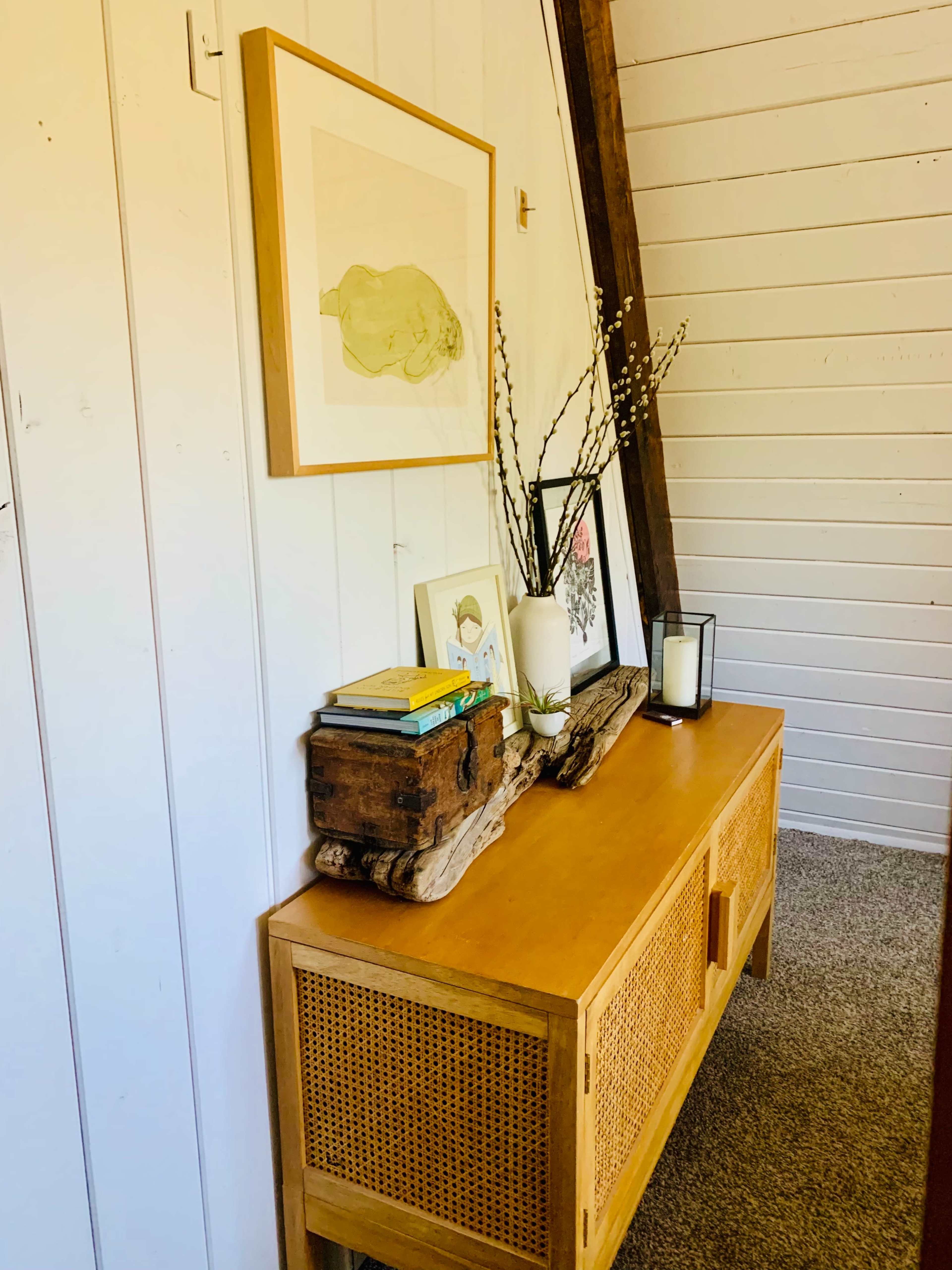 The image shows a wooden console table adorned with books, a small framed photo, and a decorative vase with branches, against a backdrop of white wooden walls.