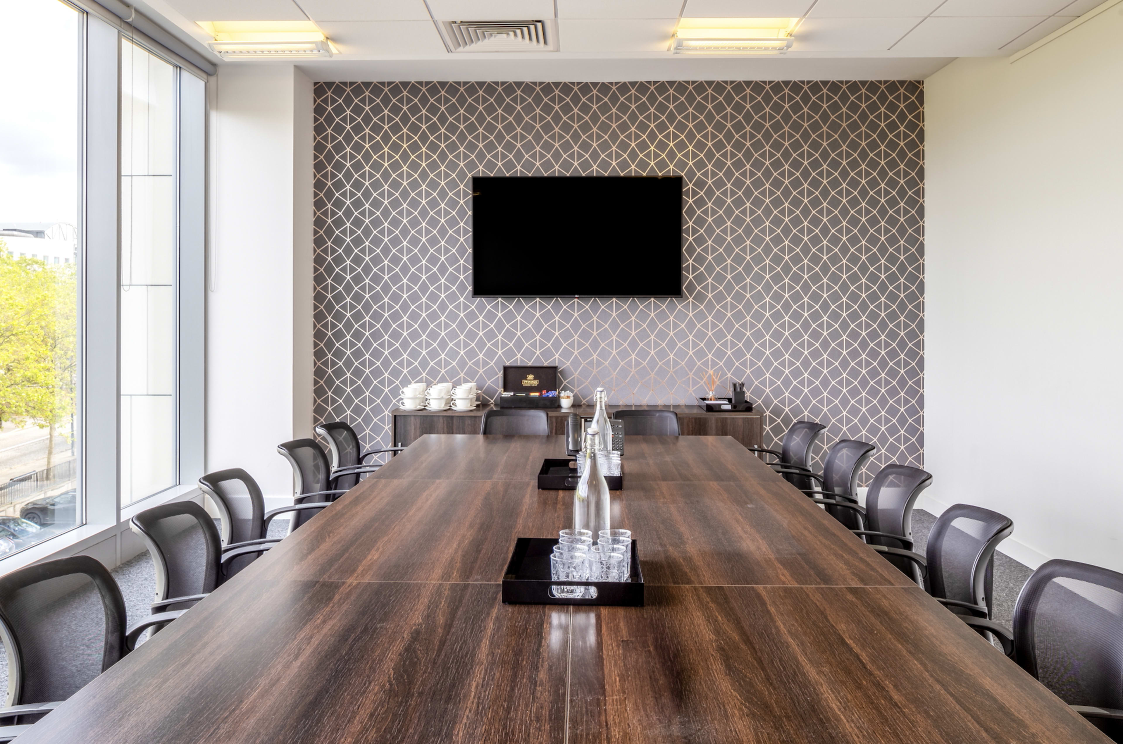 A long conference table is set in a modern meeting room with a patterned accent wall and a large screen mounted above.