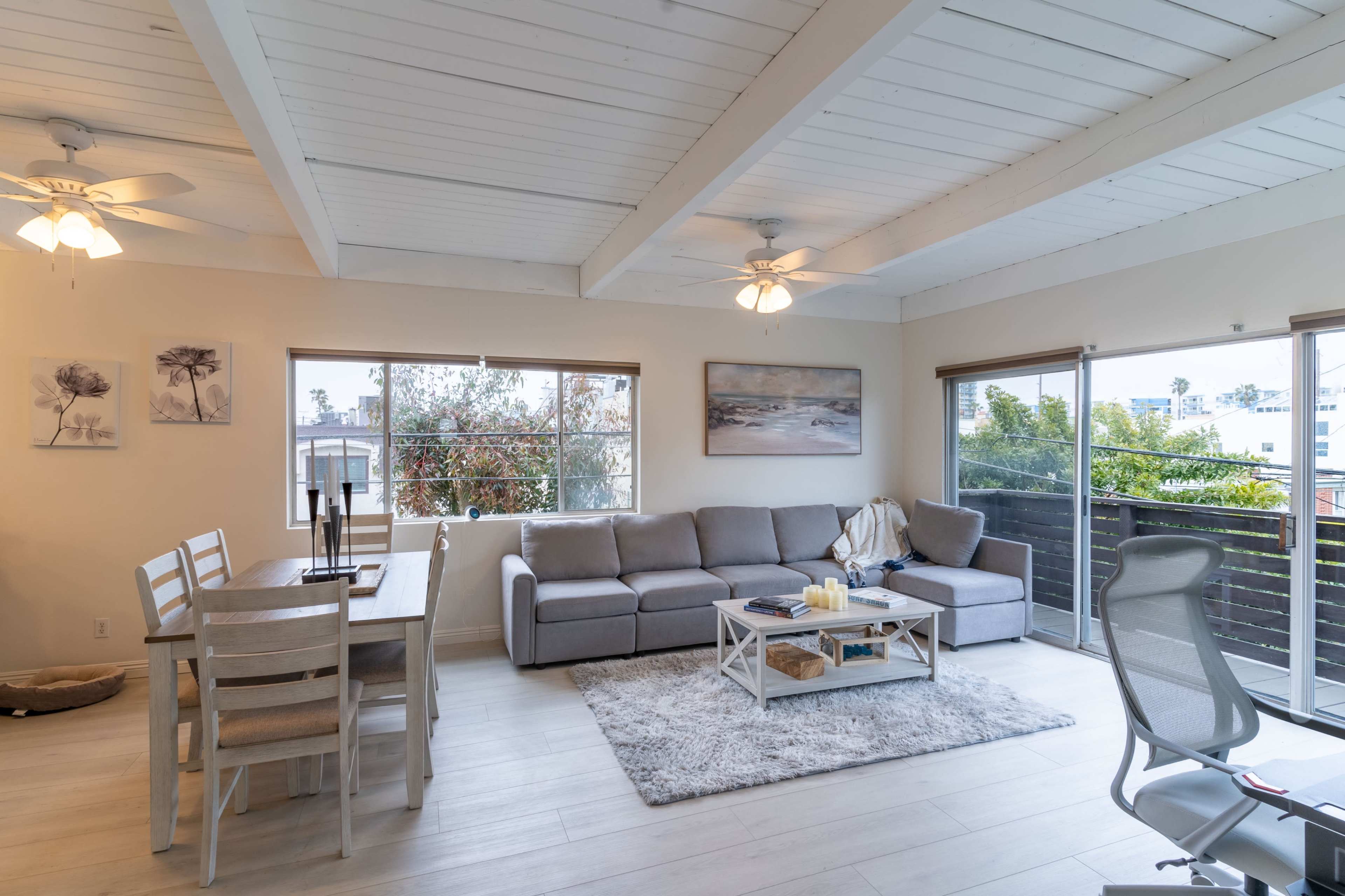 The image shows a living room and dining area with a gray sectional sofa, a light wooden table, and large windows providing natural light.