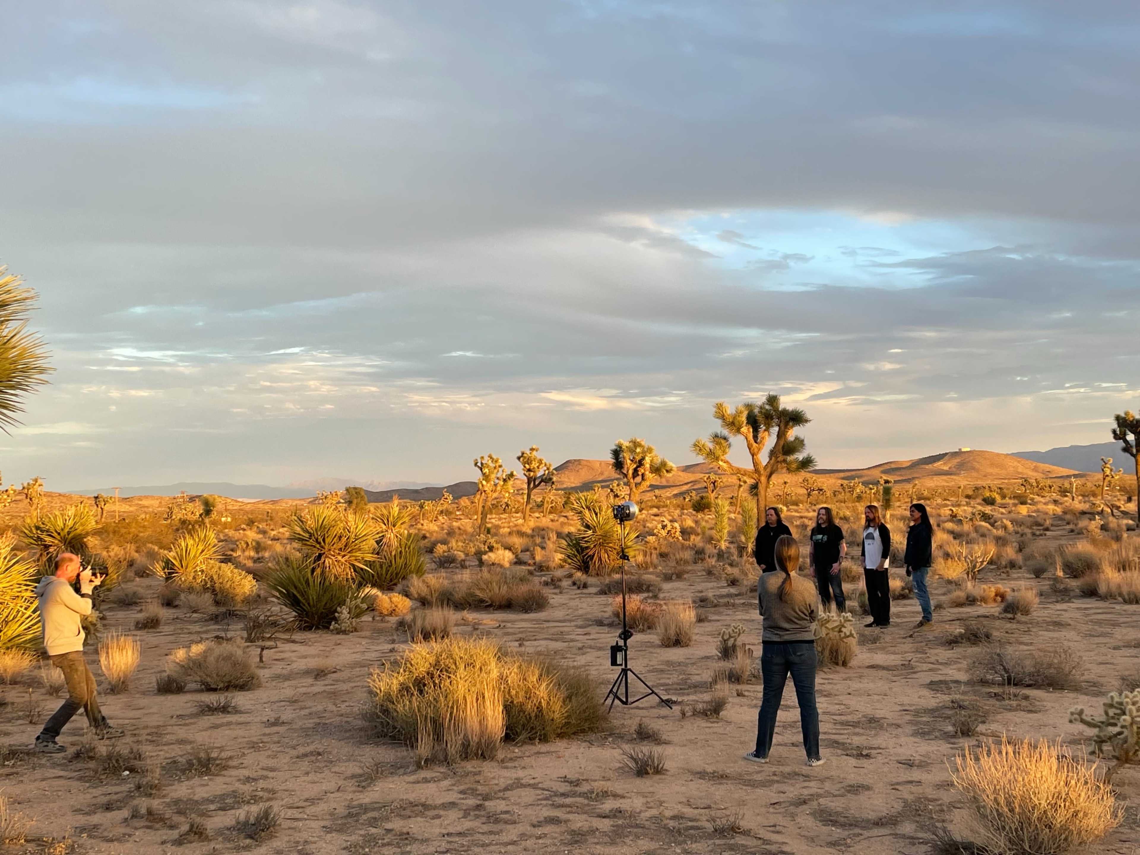 A photographer captures a group of five people posing in a desert landscape with Joshua trees and mountains in the background during sunset.