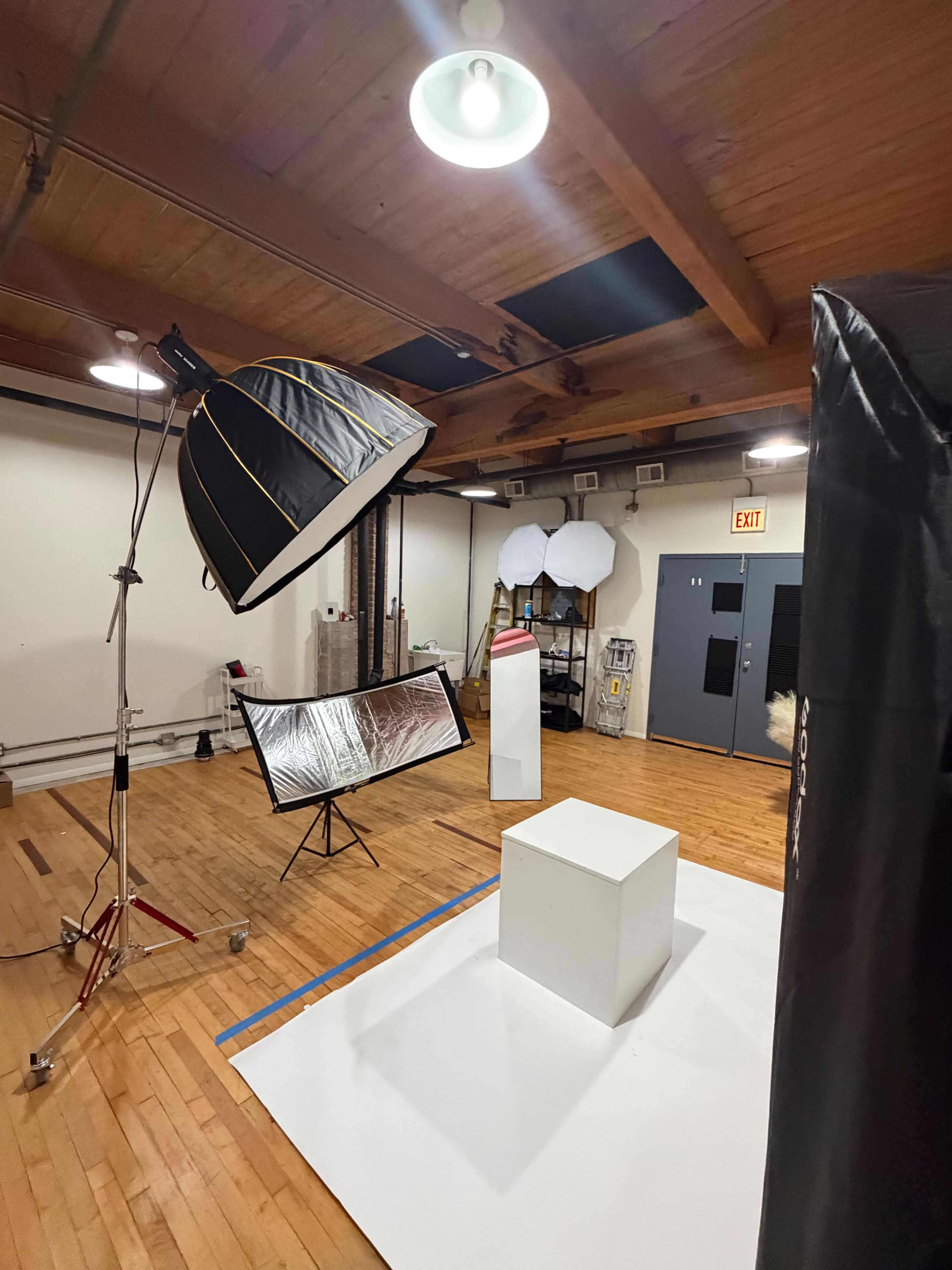 A photography studio features a large umbrella light, a reflective screen, and a white pedestal in the center of a wooden-floored space.