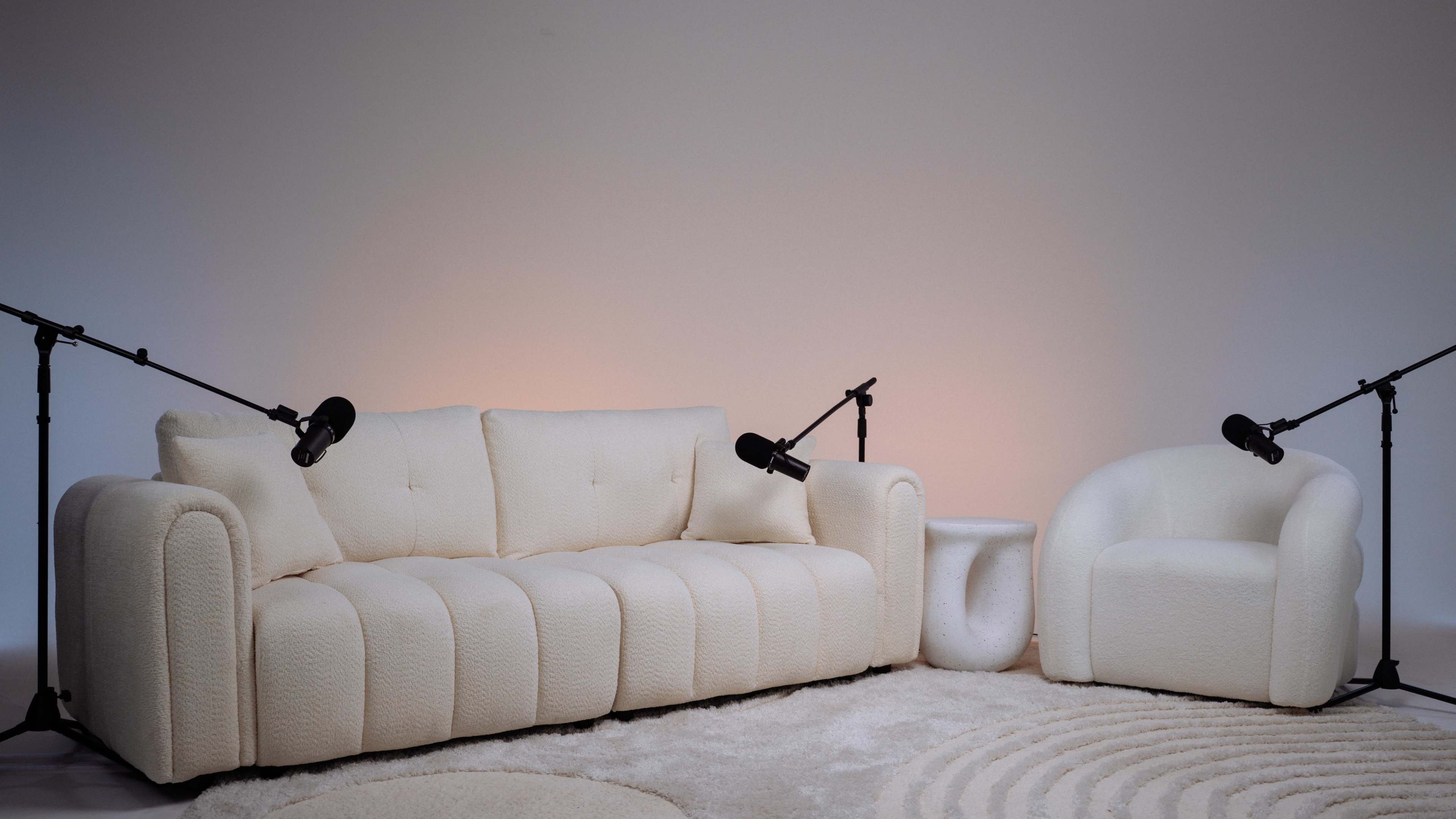The image features a white, plush sofa and a matching armchair positioned on a circular rug, with microphones attached to stands on either side.