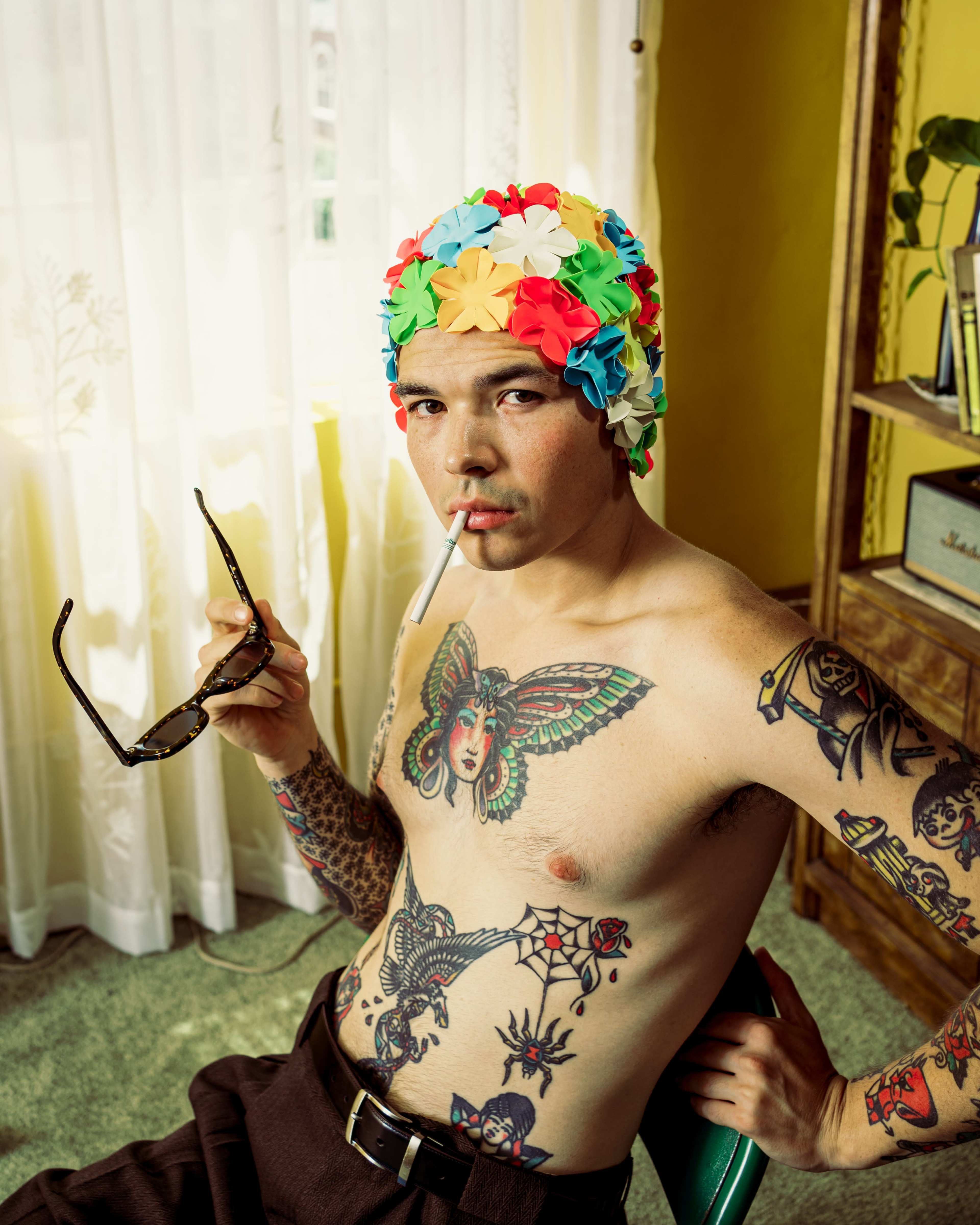 A shirtless man with tattoos sits in a chair, wearing a colorful swim cap and holding sunglasses and a cigarette.