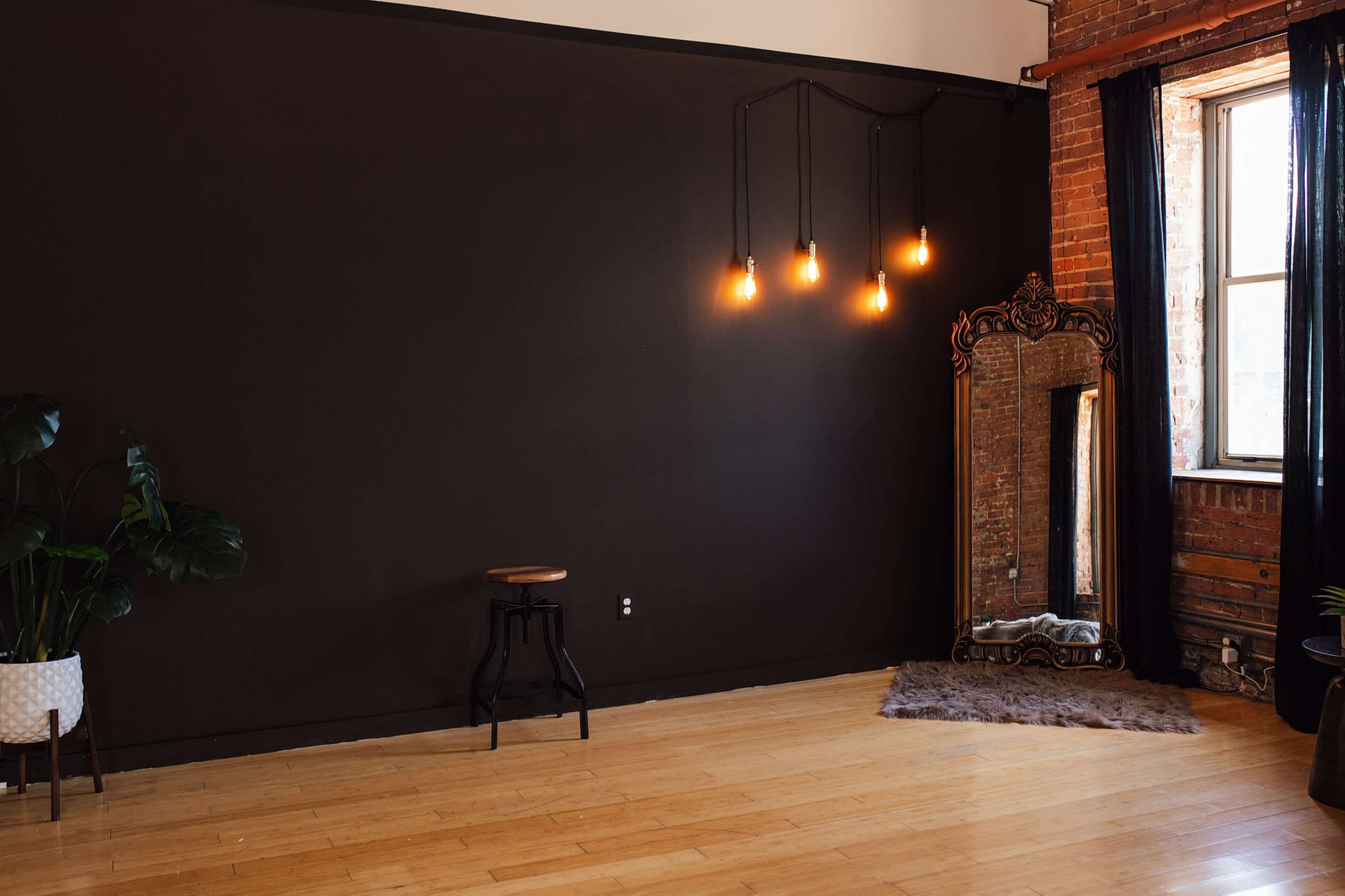 The image shows a spacious room with a dark wall, wooden floor, a small stool, a large ornate mirror, and three hanging light bulbs.