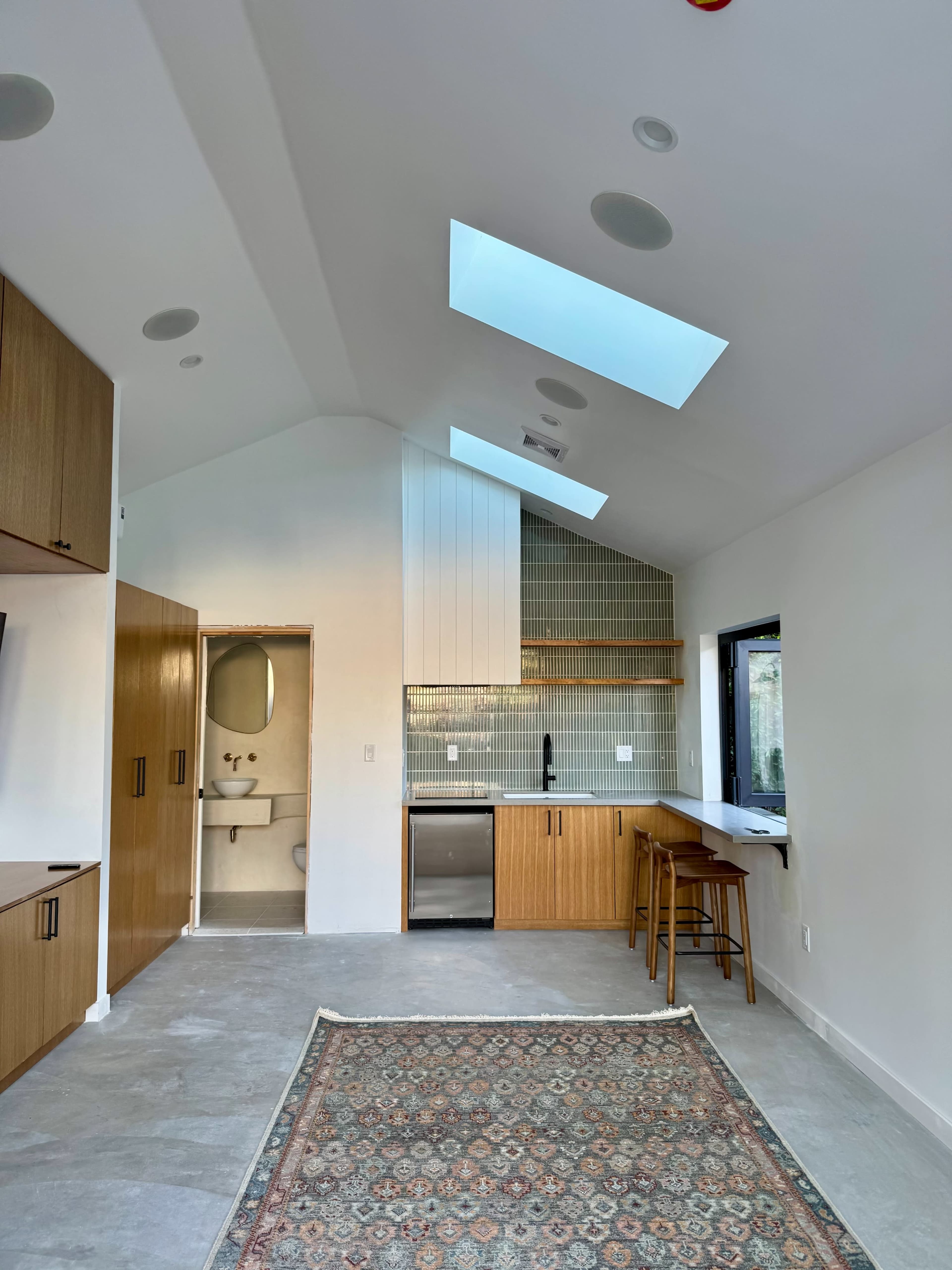 The image shows a modern kitchen and living area featuring skylights, wooden cabinetry, and a small dining space with a rug on the floor.