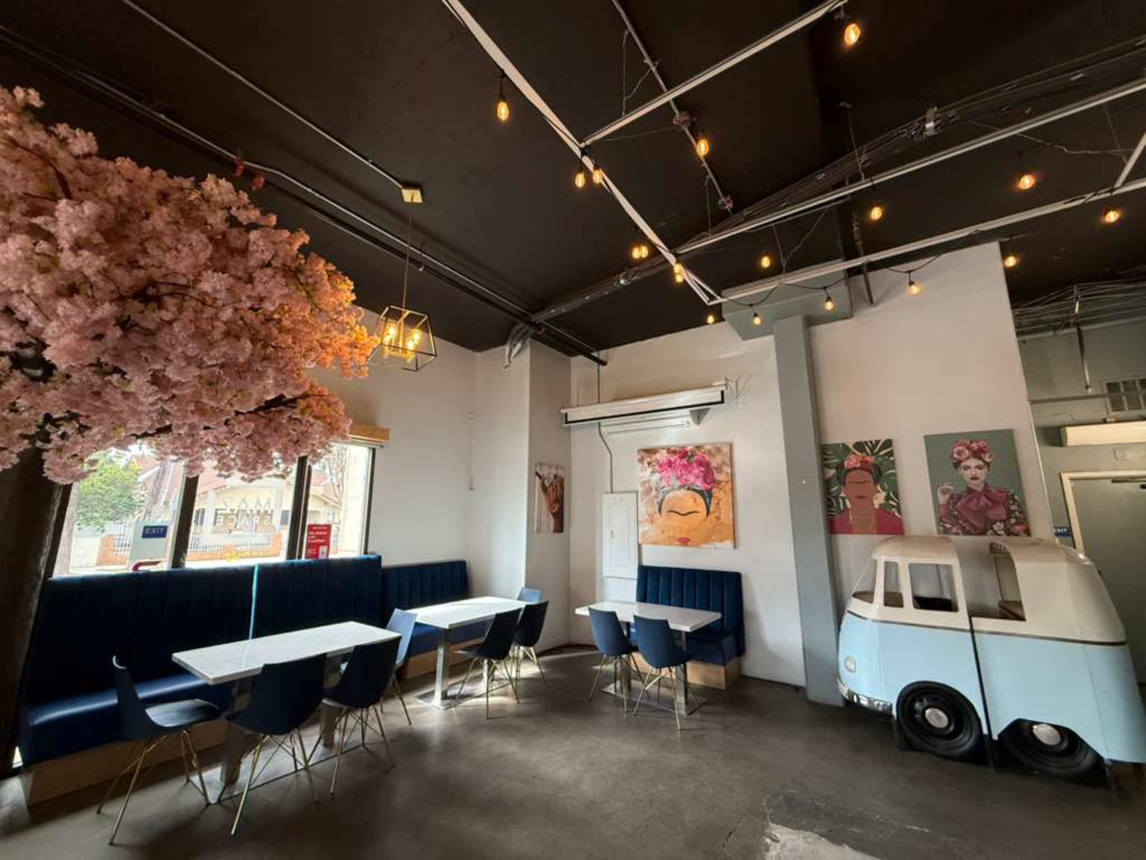 The image shows a modern cafe interior featuring blue seating, pink flowering trees, and artistic wall decorations, with a whimsical white car-shaped booth.