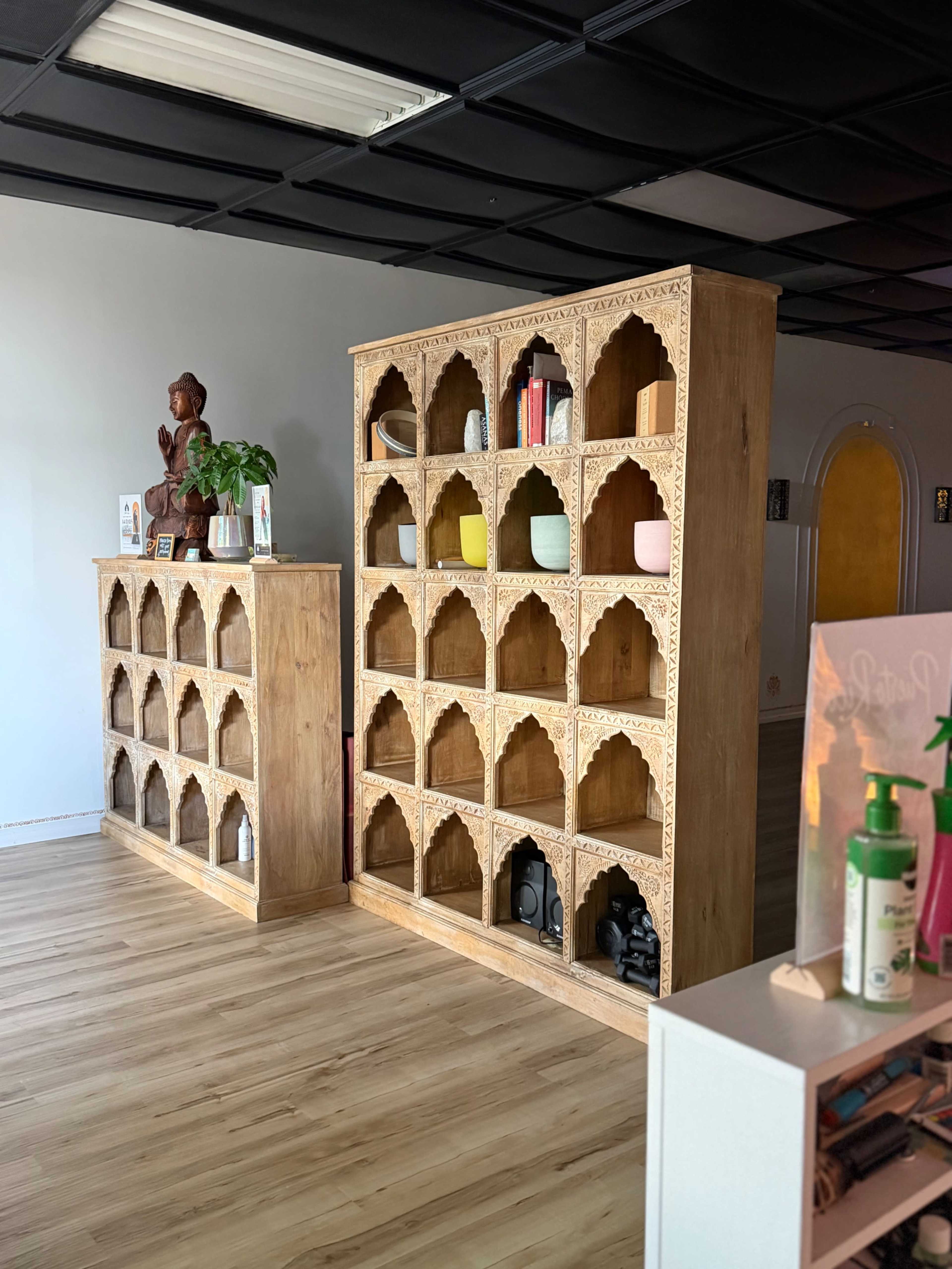 The image shows two wooden shelving units with decorative arches, displaying various items including books, plants, and decorative statues, in a minimal interior space.