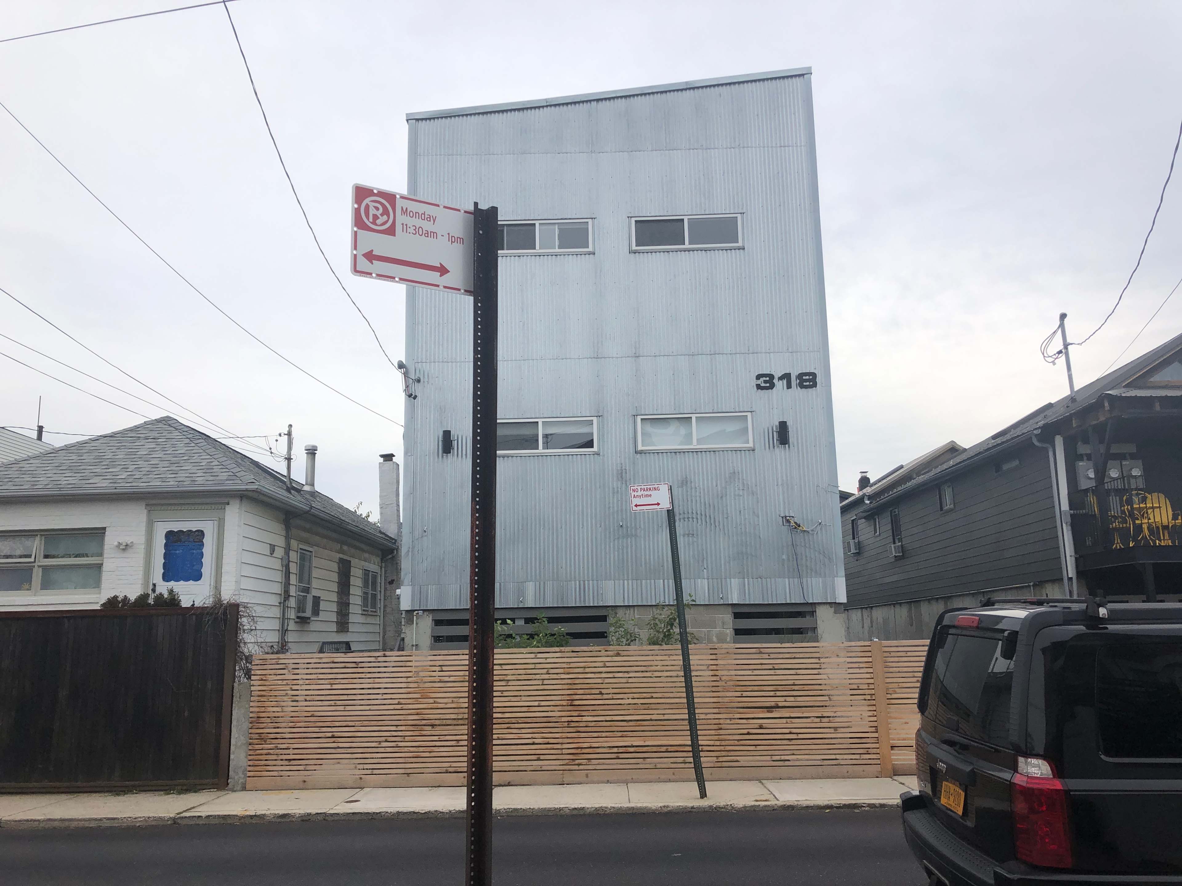 The image shows a three-story, corrugated metal building numbered 318, located on a street with a parking restriction sign and a wooden fence nearby.