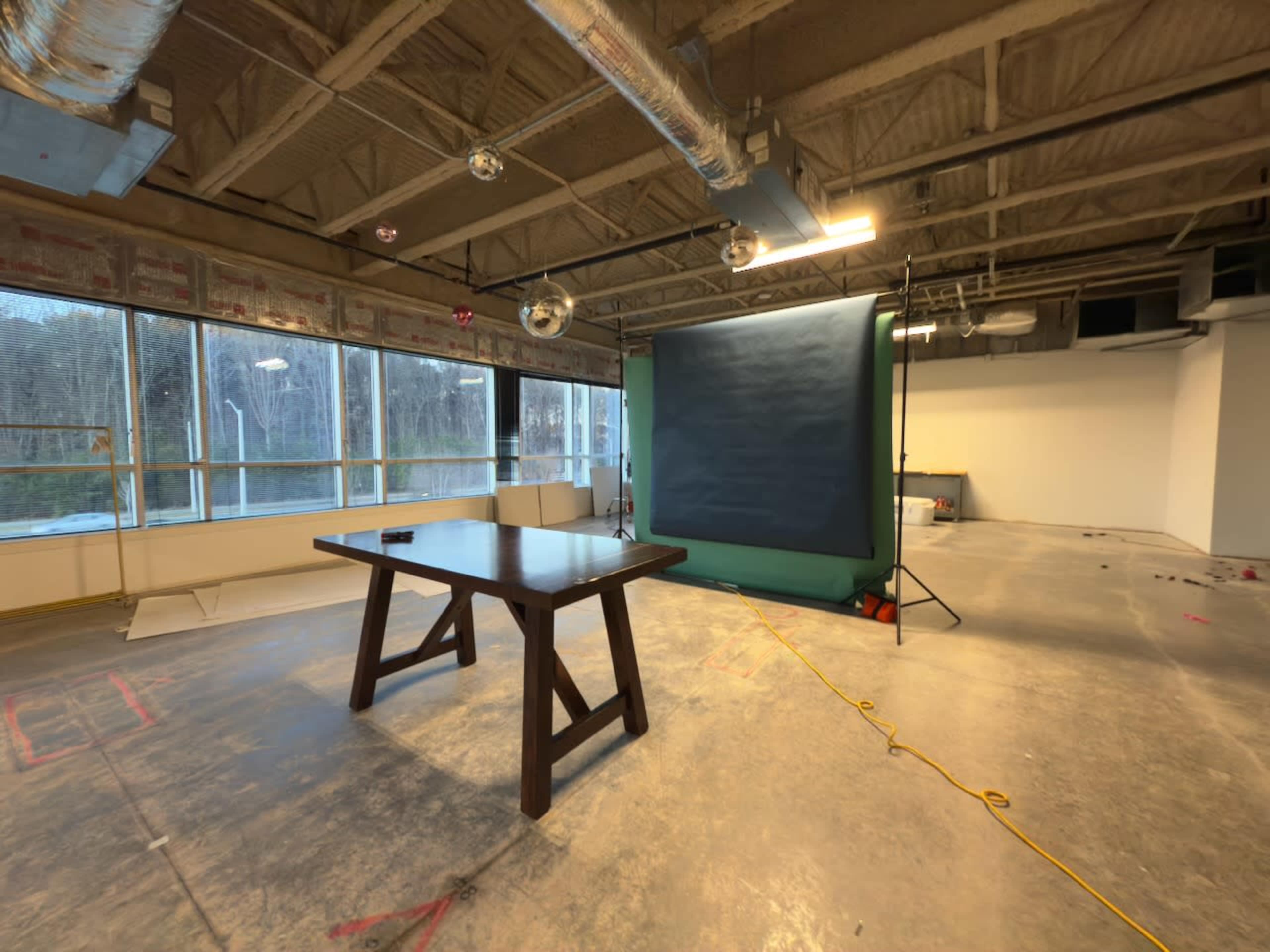 The image shows an empty room with a large window, a wooden table in the center, and a backdrop setup in the corner.