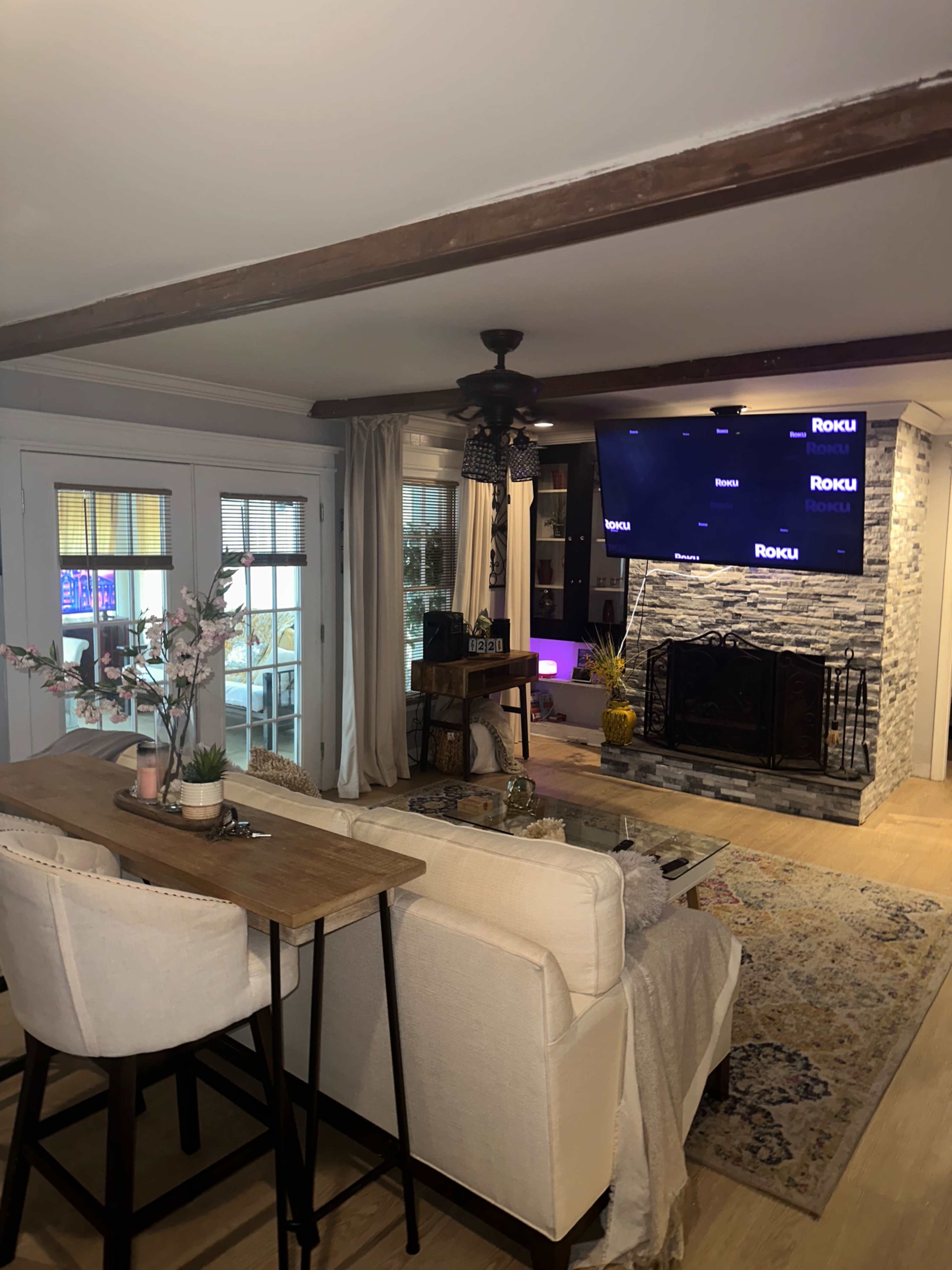 A modern living room with a stone fireplace, a television displaying the Roku interface, and a seating area featuring a sofa and a small dining table.