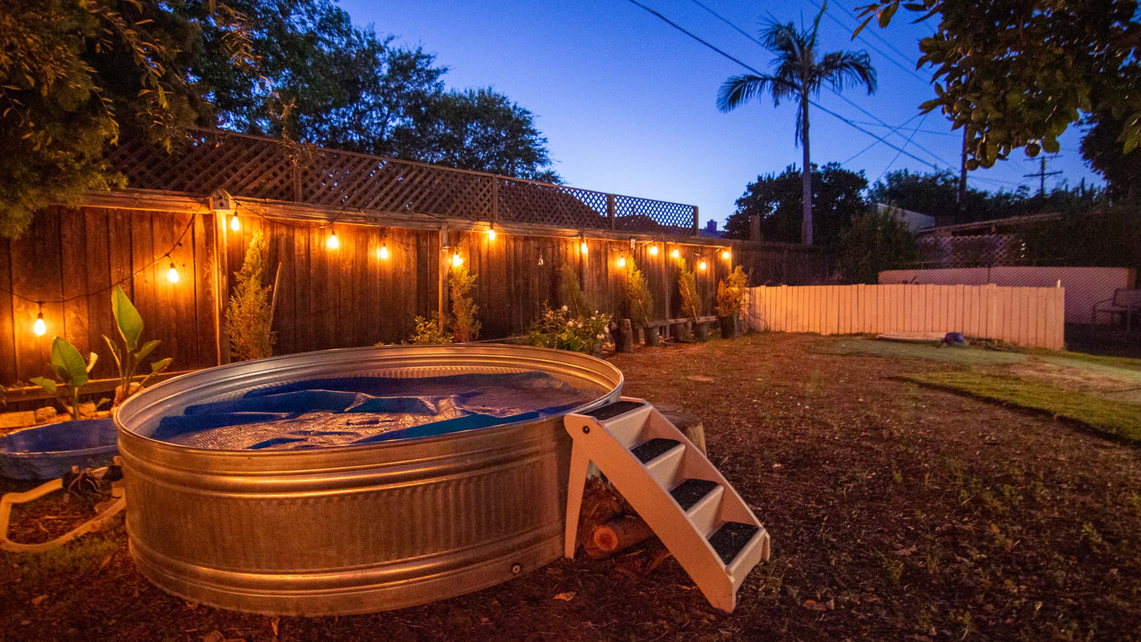 A metal stock tank pool is set up in a backyard illuminated by string lights against a twilight sky.