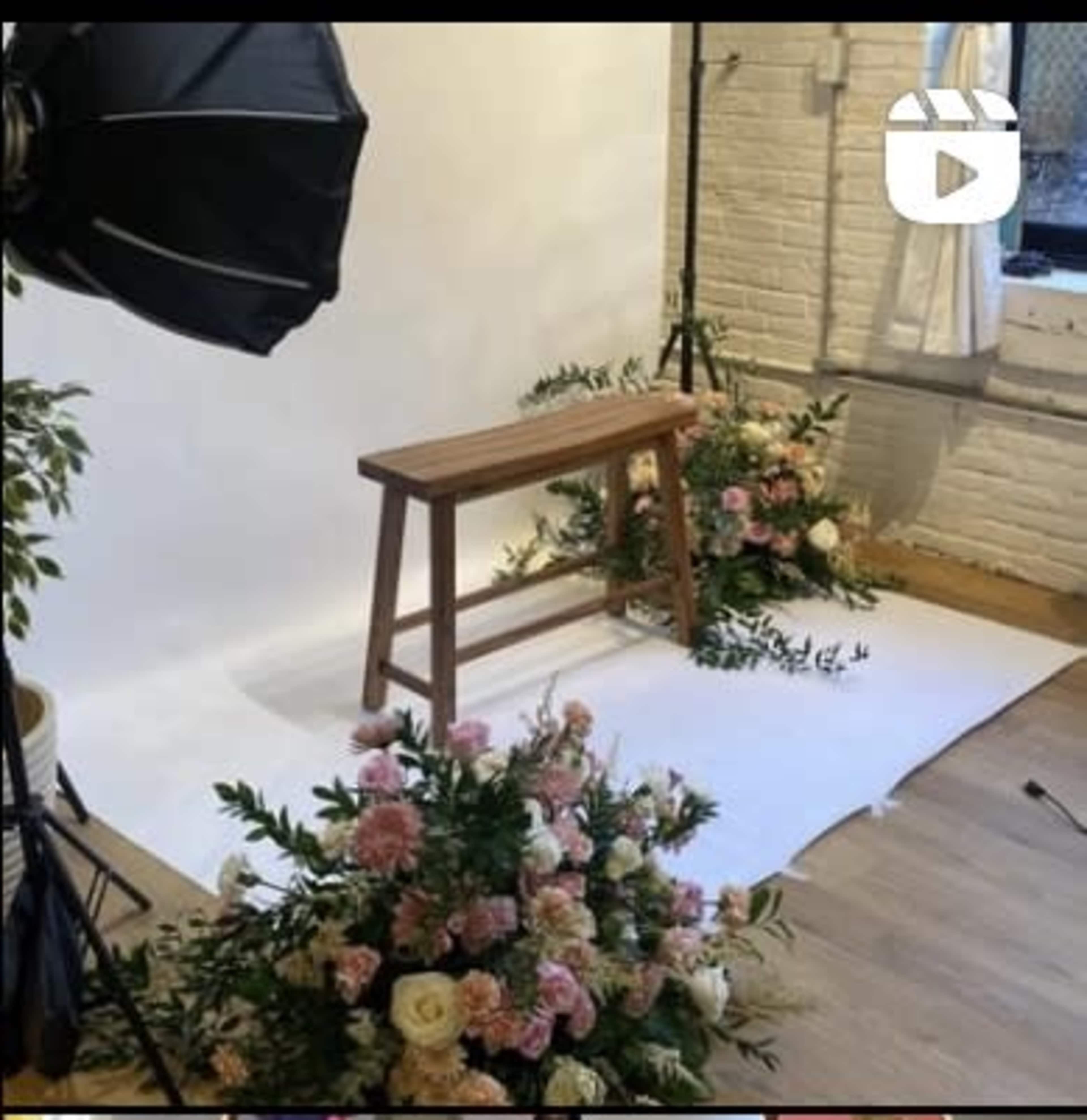 The image features a wooden stool positioned in front of a white backdrop, surrounded by colorful floral arrangements on the floor.
