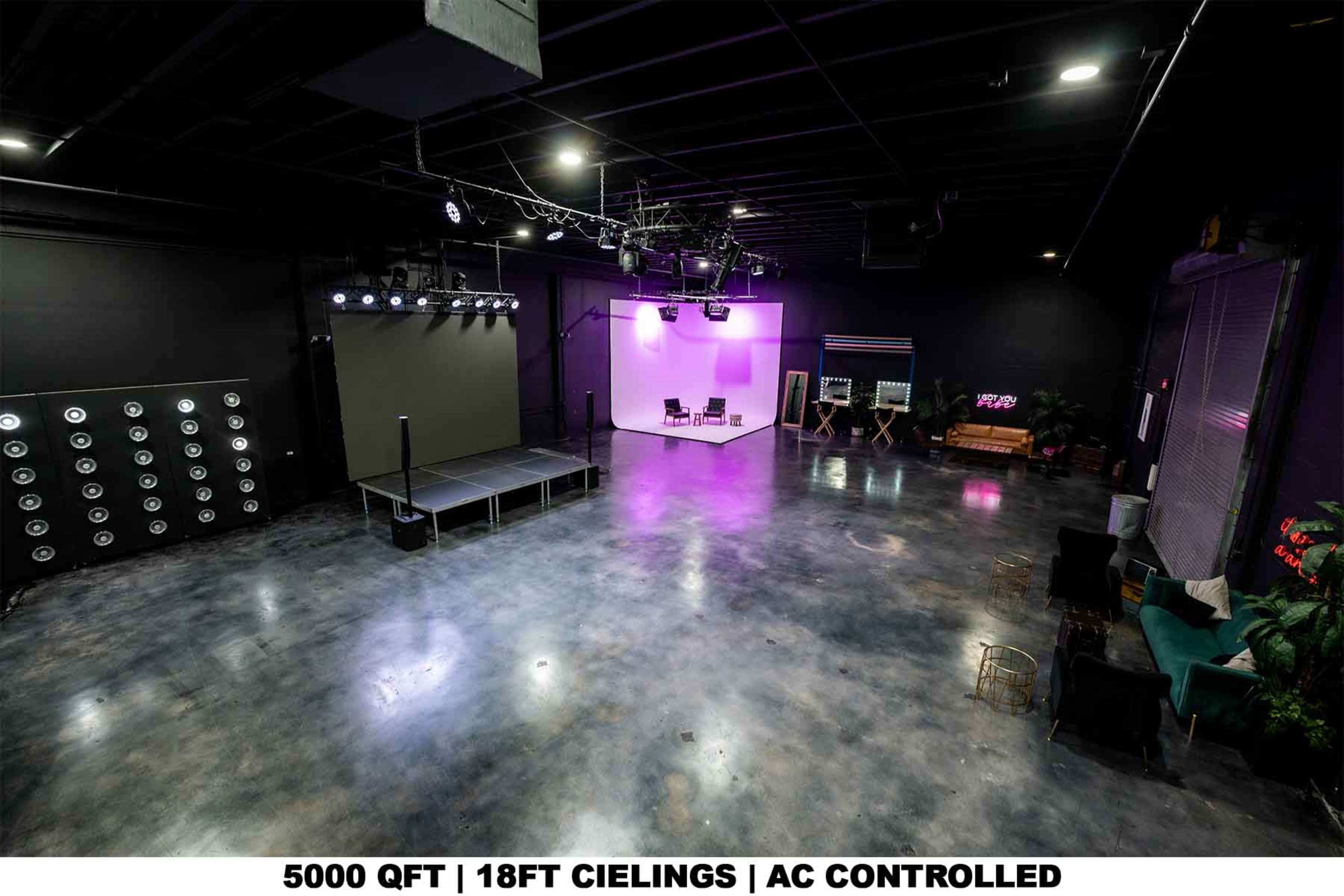 The image depicts a spacious, empty studio with a polished concrete floor and black walls, featuring staging equipment and colored lighting setup.