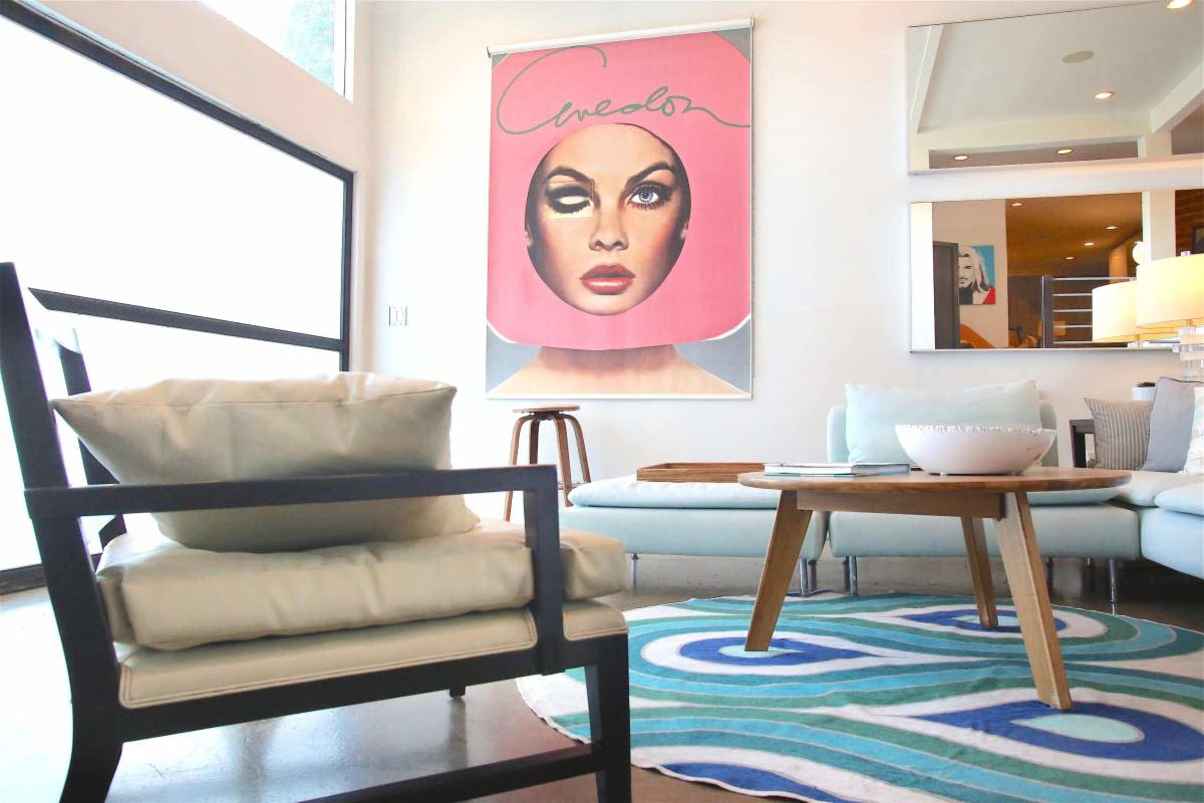 The image shows a modern living room with a large artwork featuring a woman's face on the wall, a small wooden stool, a light blue couch, and a round wooden coffee table on a patterned rug.