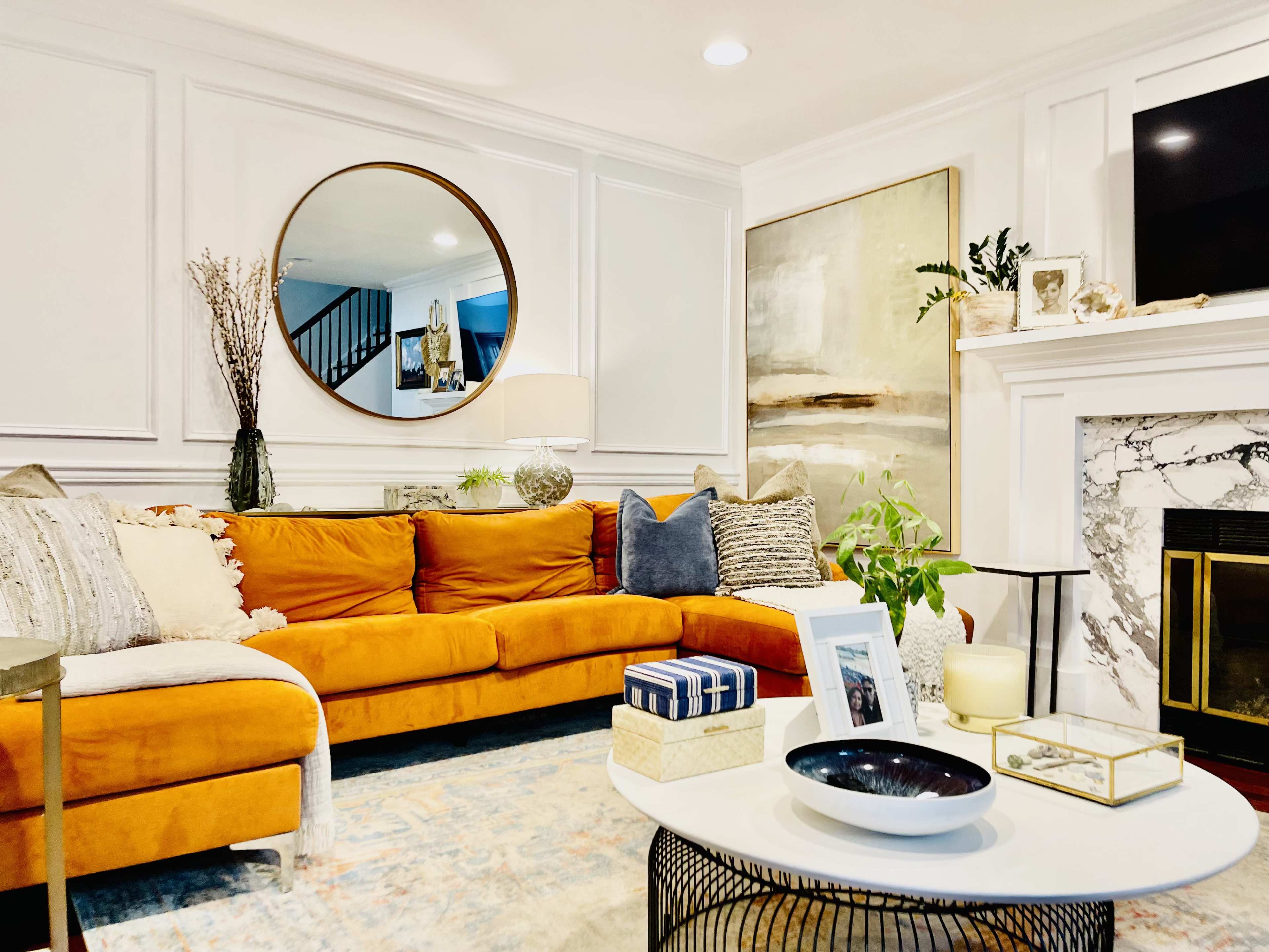 A modern living room features a large orange sectional sofa, a round mirror above a decorative fireplace, and various art pieces and plants arranged thoughtfully throughout the space.