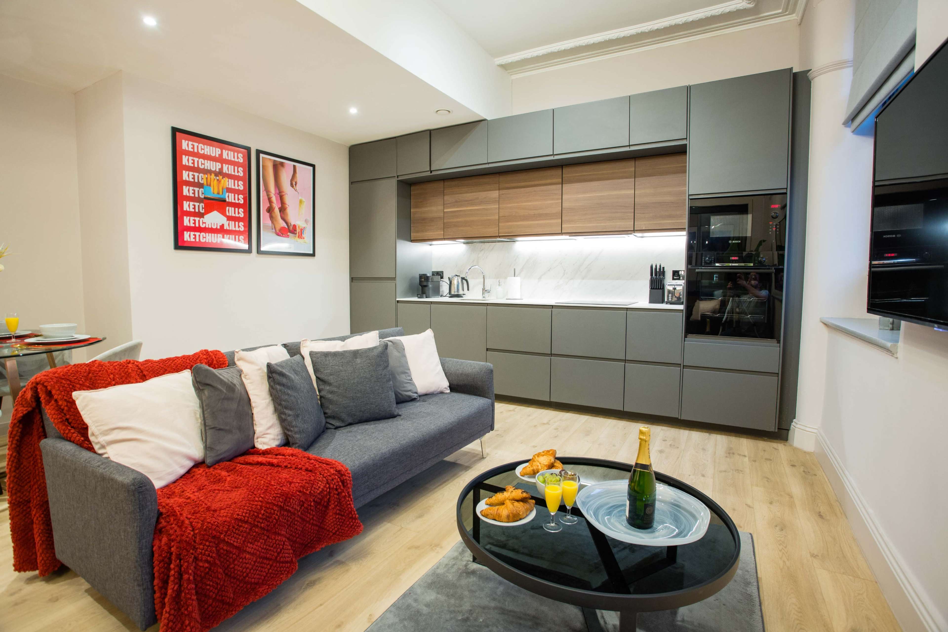A modern living room features a gray sofa with colorful cushions, a round coffee table with snacks and drinks, and a sleek kitchen area with dark cabinets and a marble backsplash.