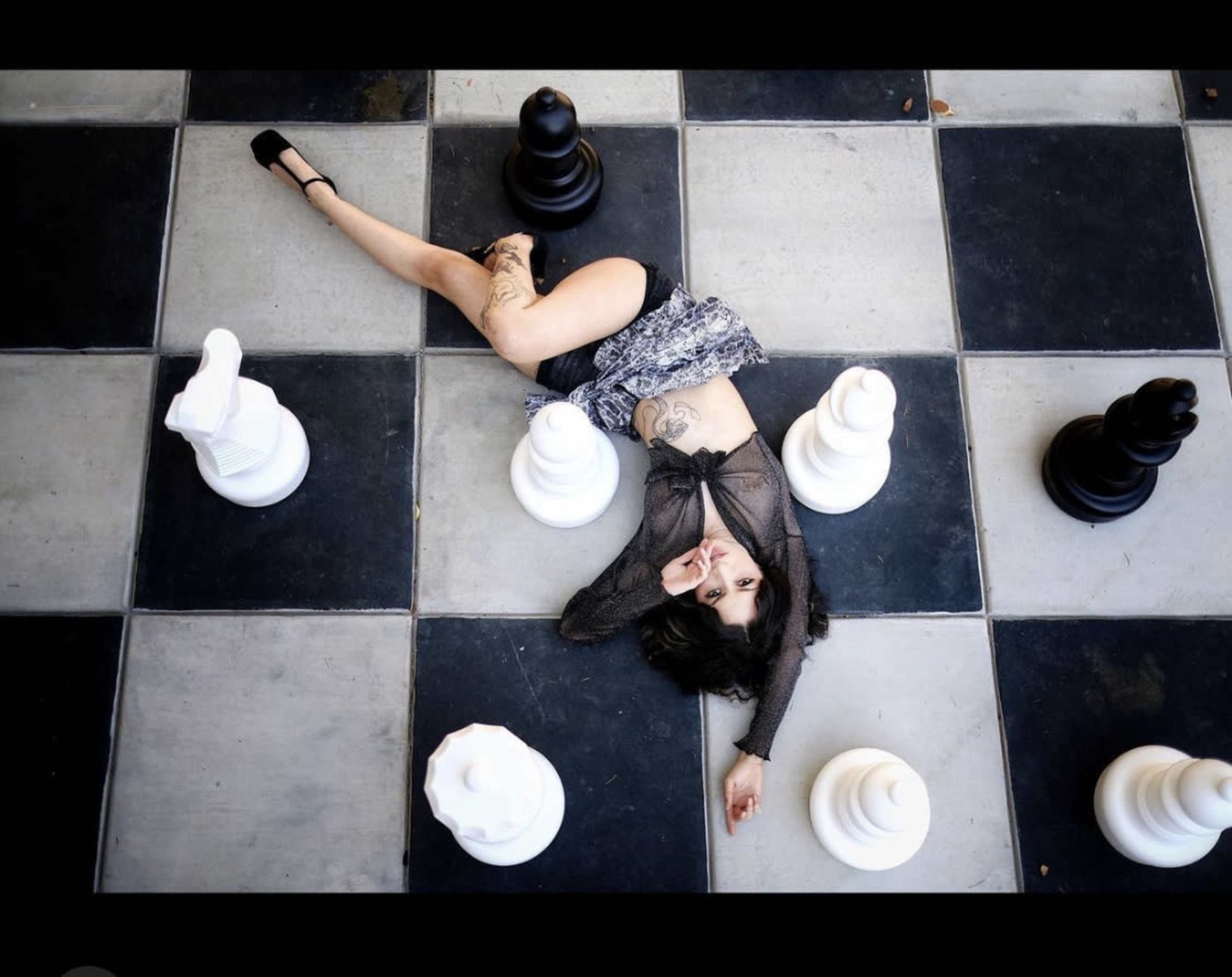 A person lies on a large checkered chessboard, surrounded by oversized black and white chess pieces.