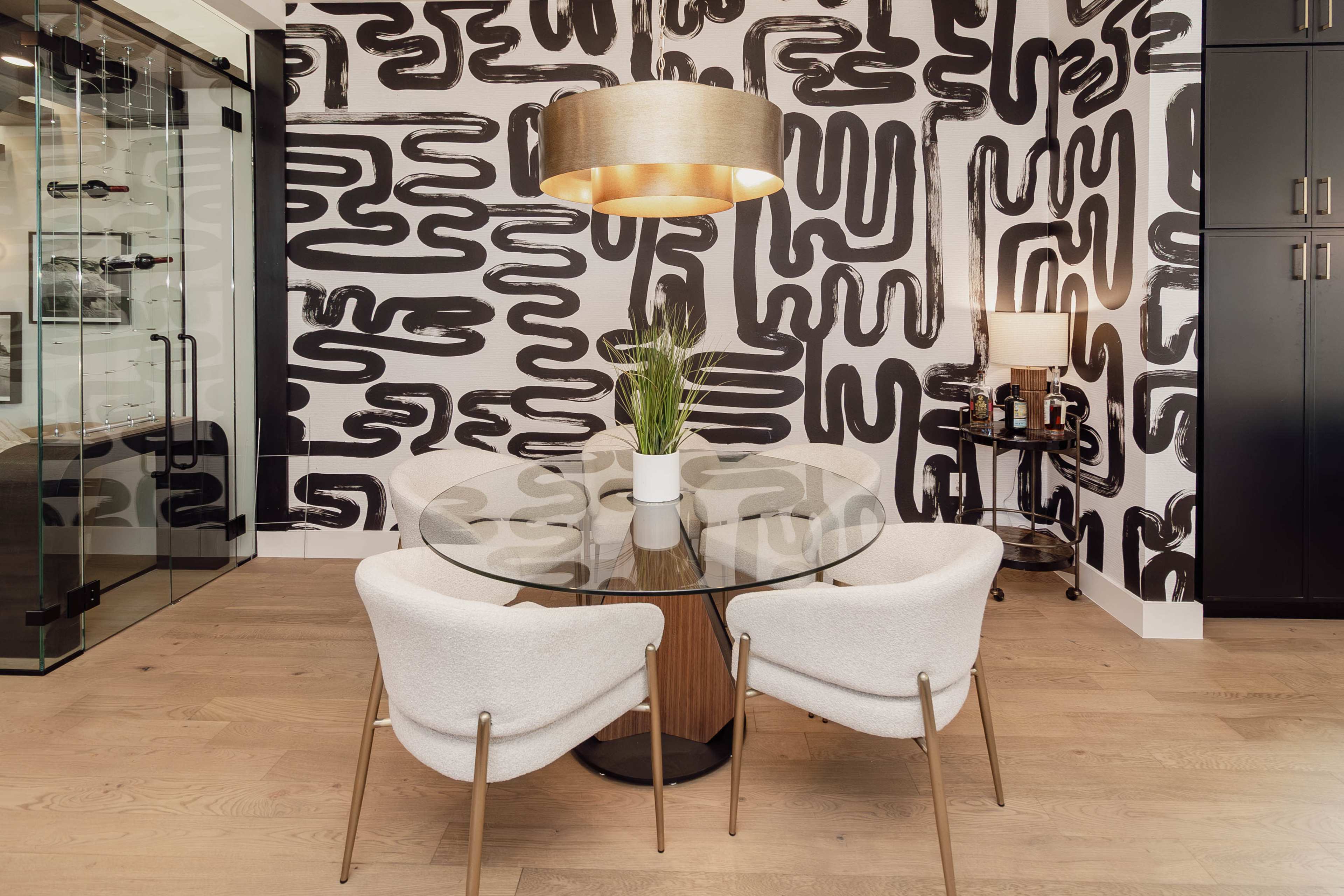 A round glass dining table with white chairs is set against a black and white abstract patterned wall, with a pendant light hanging above and a plant centerpiece.