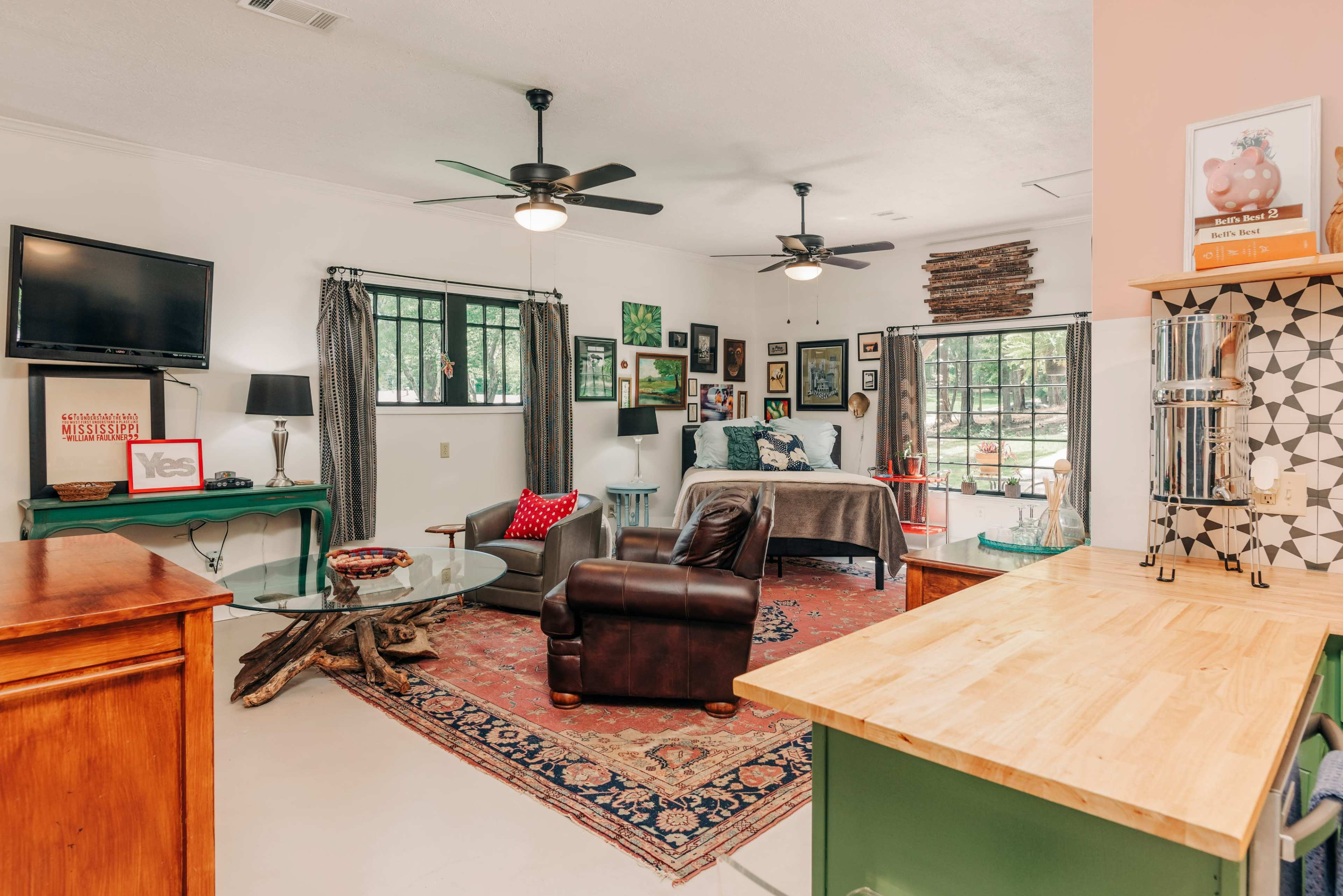 A spacious room features a living area with a brown leather chair, a dining table, and large windows that overlook an outdoor view, with various artworks displayed on the walls.