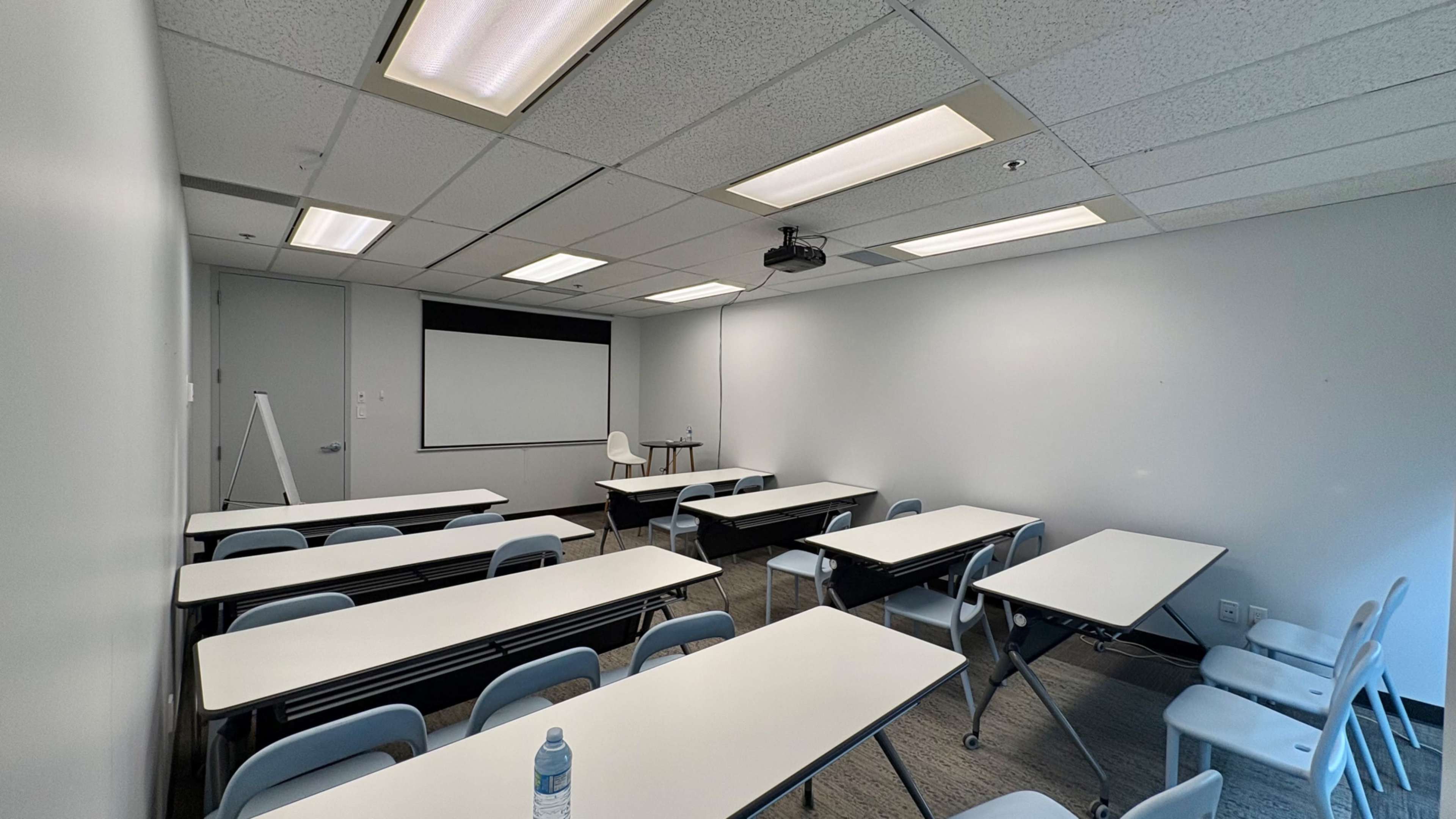 A classroom is set up with a projector, whiteboard, and several tables and chairs arranged in a simple layout.