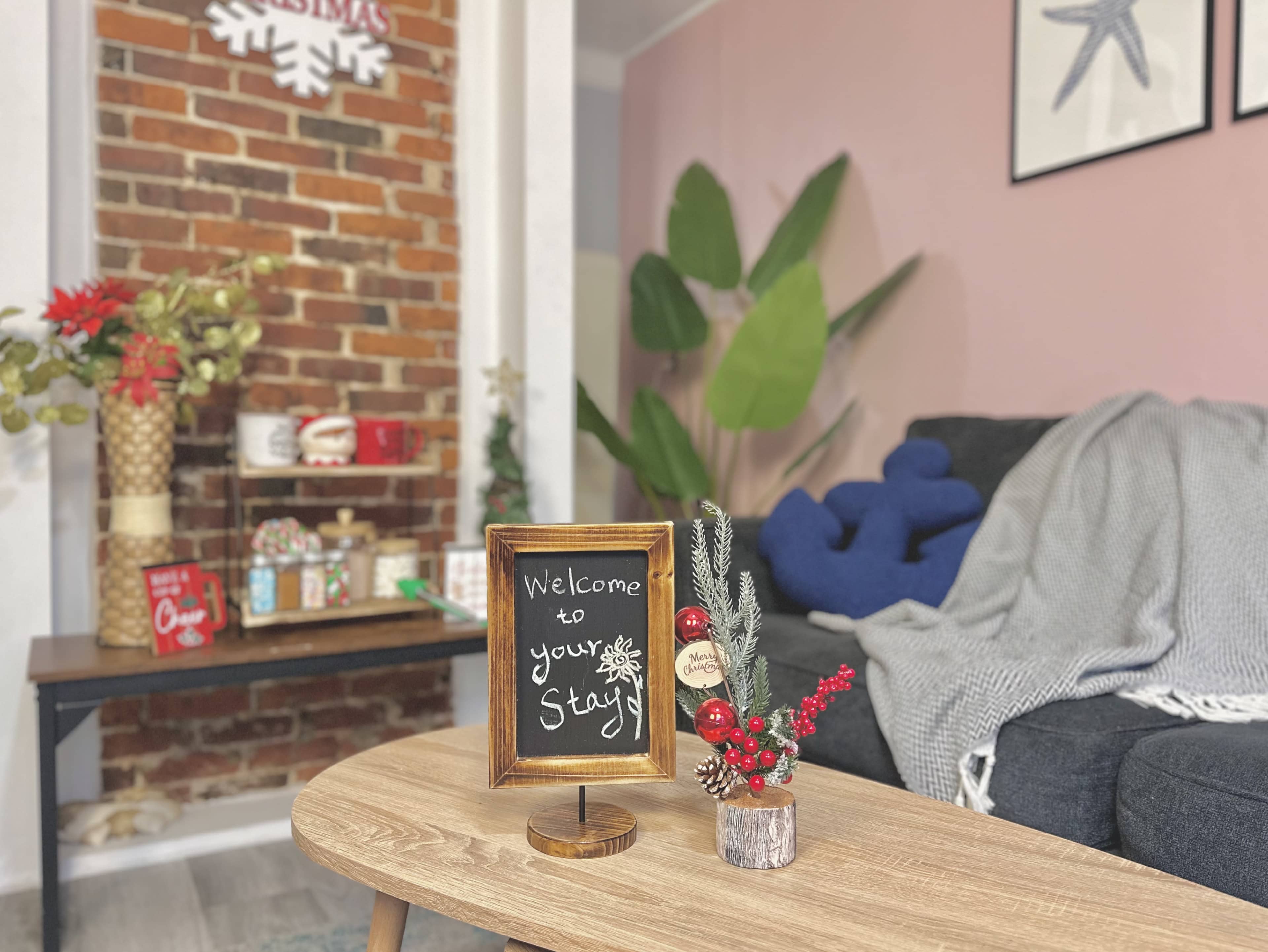 A cozy living room features a chalkboard sign welcoming guests, a small decorative plant, and a couch near a brick wall adorned with holiday decorations.