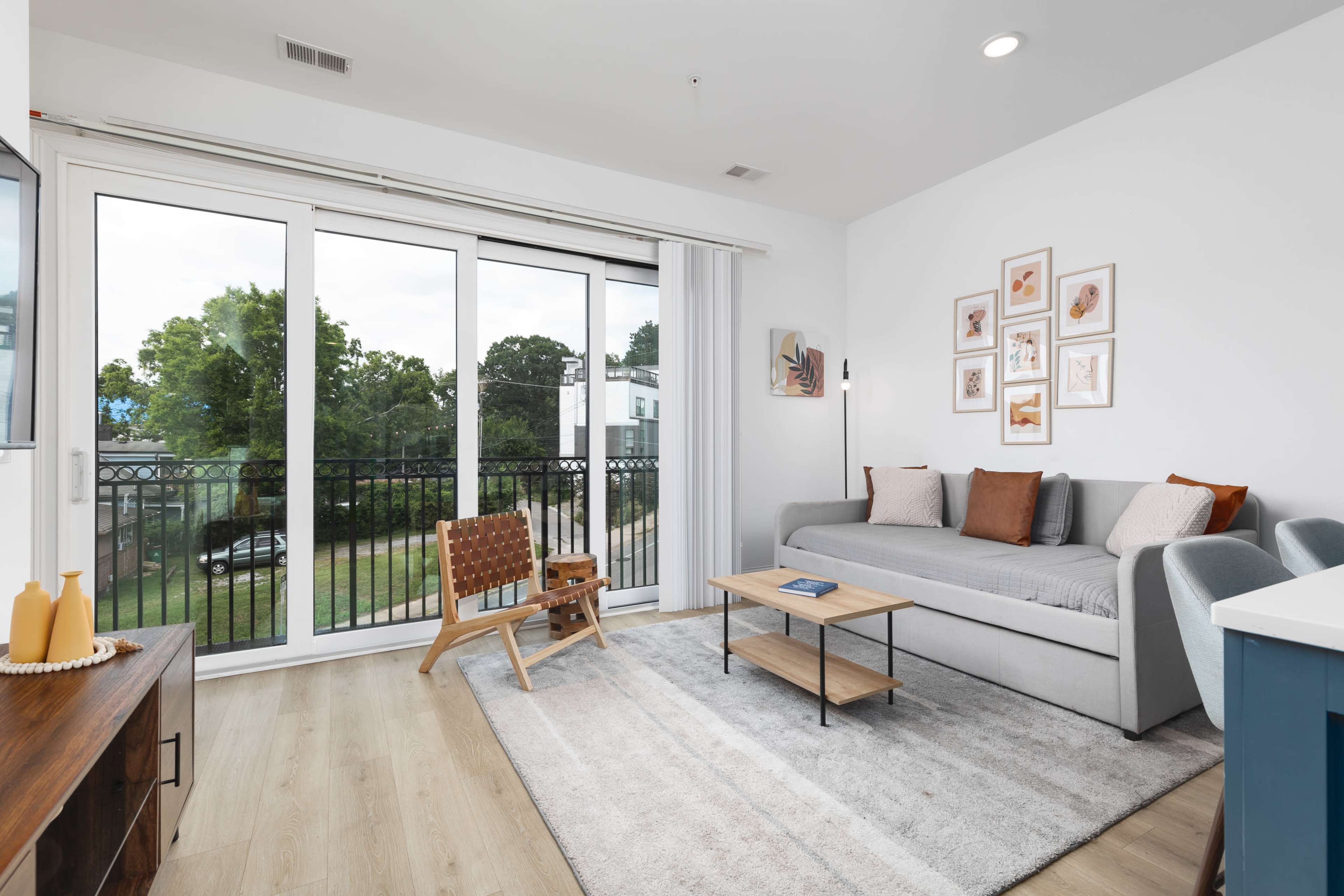 The image shows a modern living room with a grey sofa, a wooden chair, a coffee table, and large windows that overlook a green space.