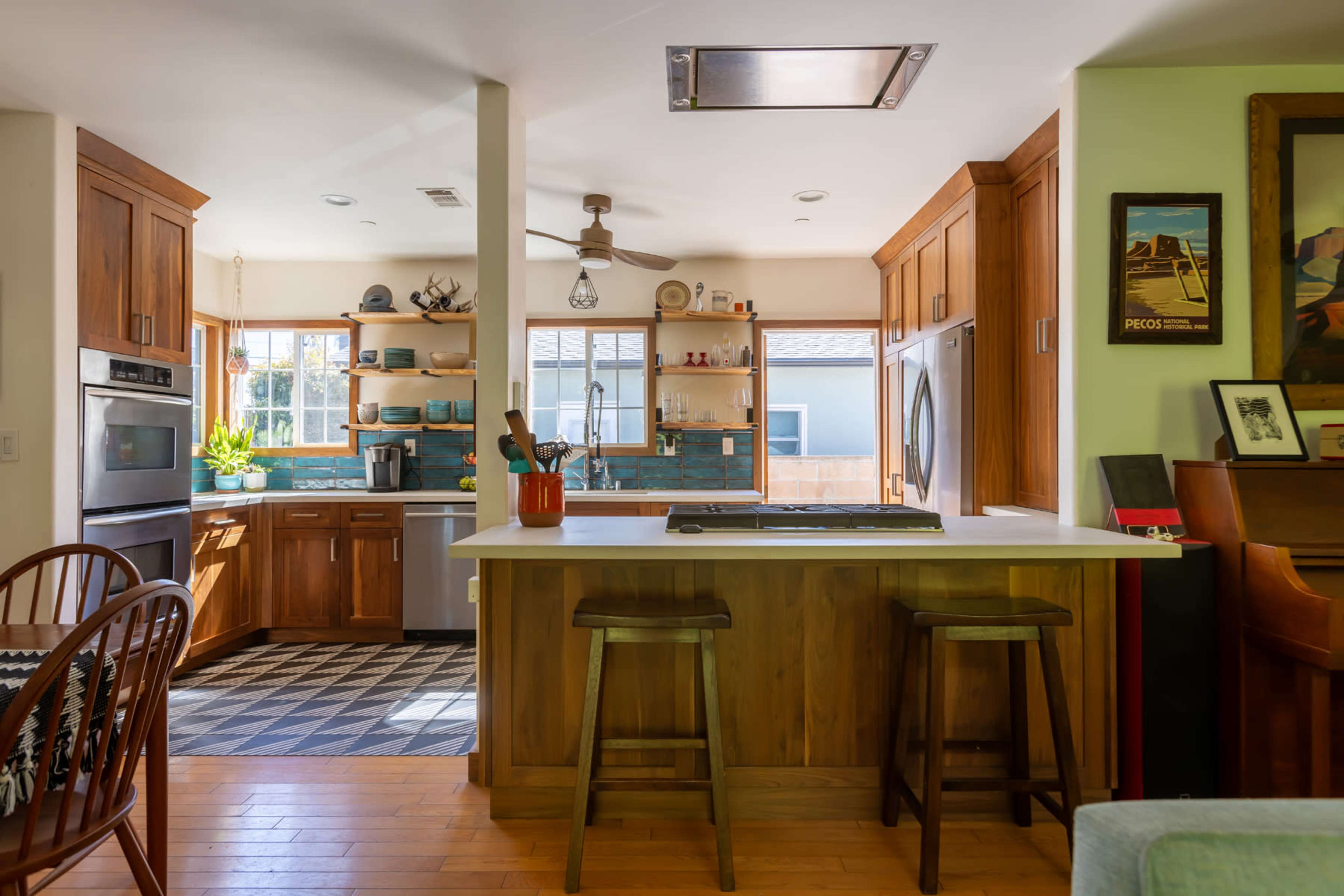 The image shows a modern kitchen with wooden cabinets, a central island with bar stools, and a blend of natural light and blue accents.