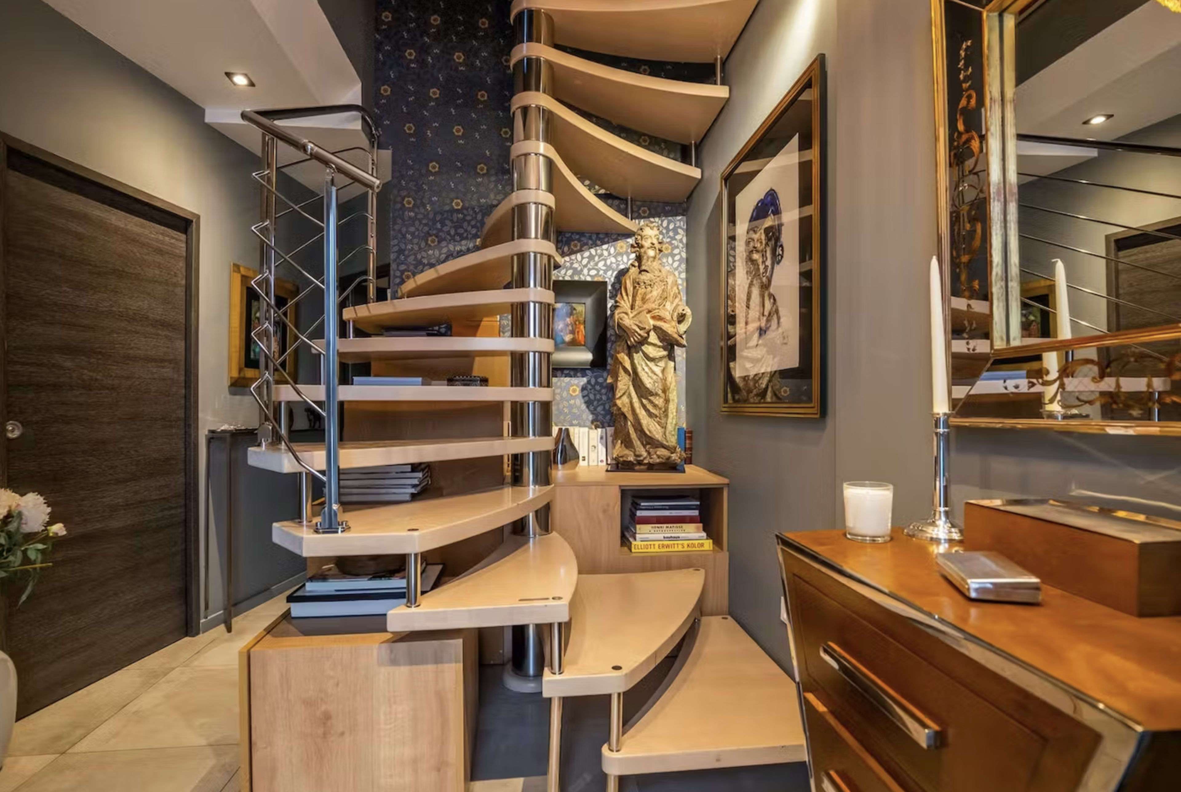 The image features a modern spiral staircase made of wood, surrounded by wall art and decorative items in a well-lit hallway.
