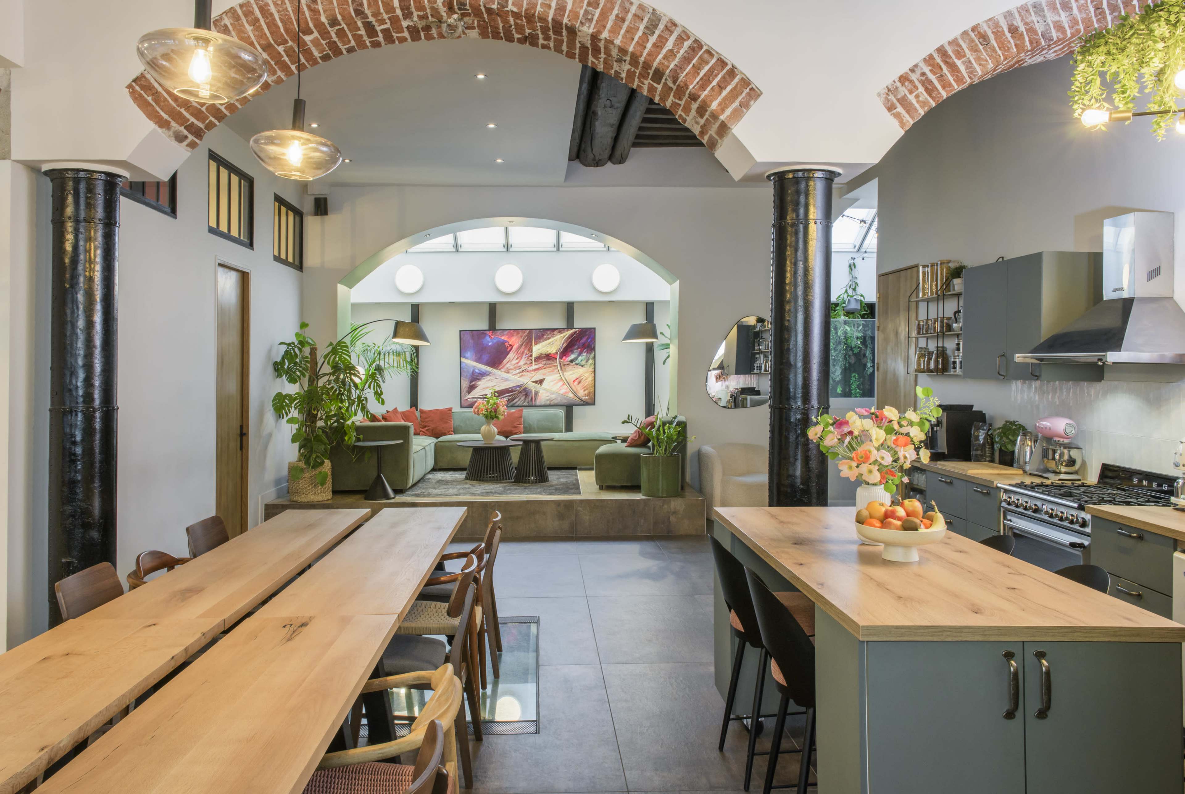 The image shows a contemporary open-concept kitchen and dining area, featuring a long wooden table, green cabinetry, and a spacious living area with a large screen and plush seating.