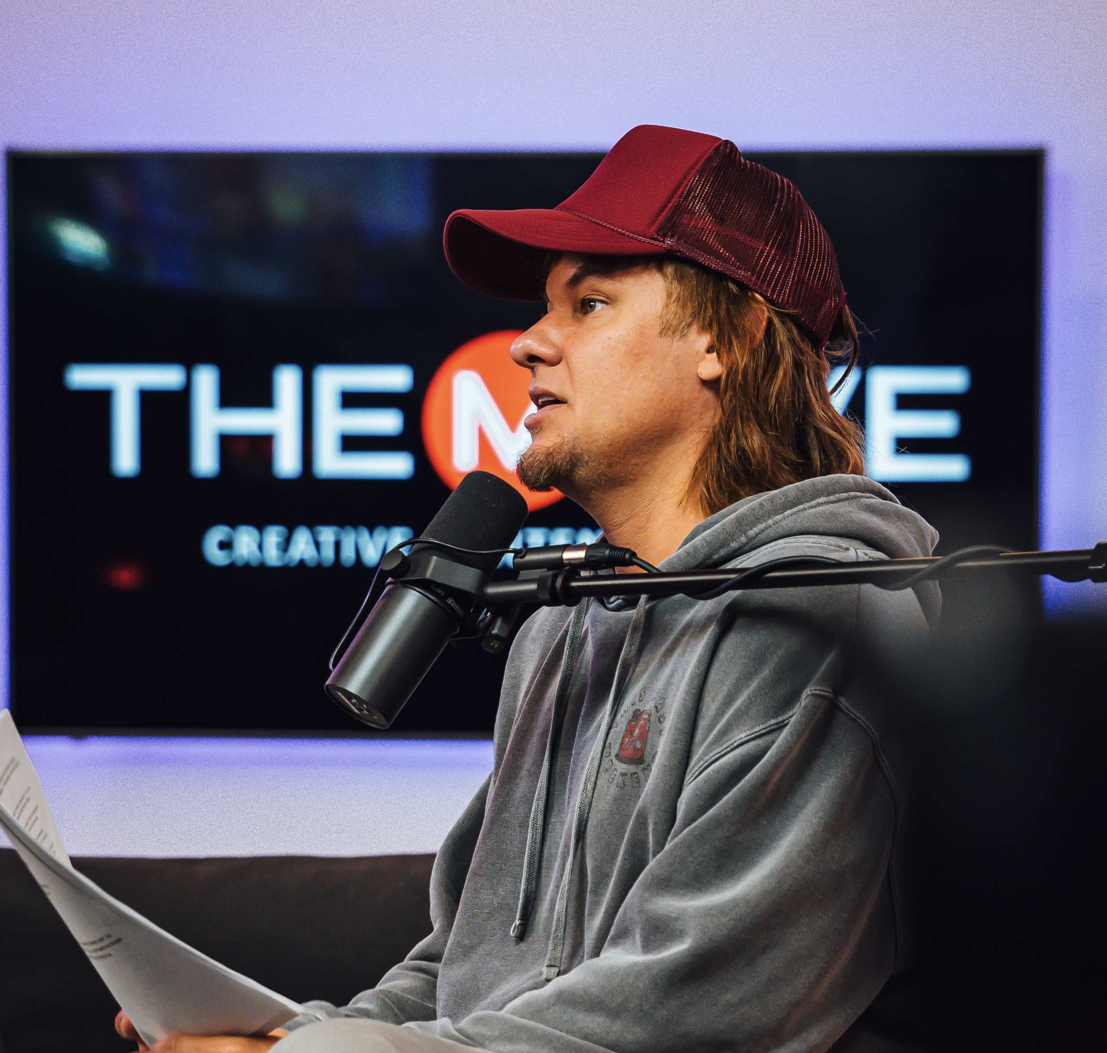 A person with long hair wearing a baseball cap and a hoodie sits in front of a microphone while looking at a sheet of paper.