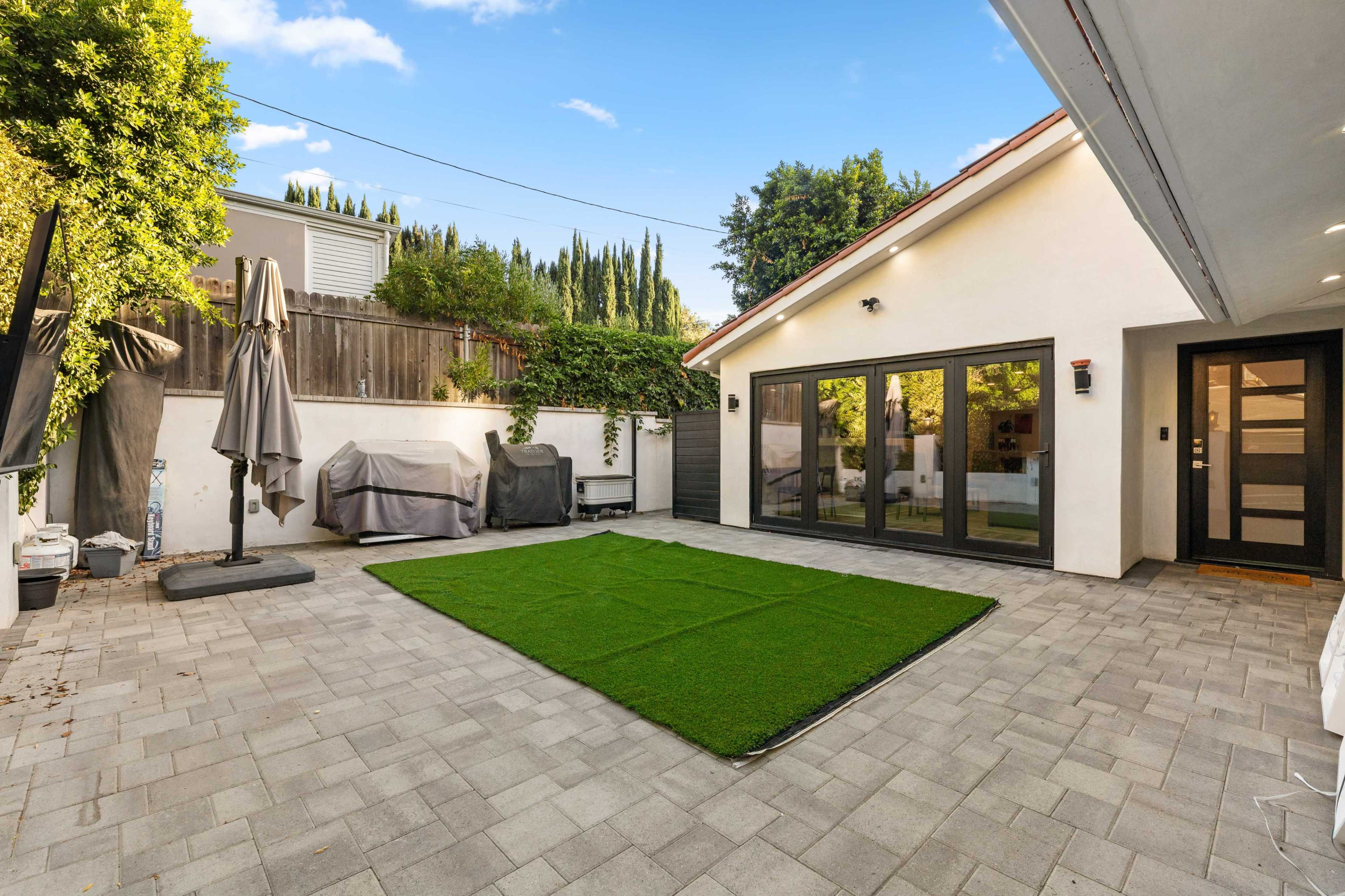 The image shows a patio area with a green artificial lawn, outdoor furniture covered with a grey tarp, and a modern house with large windows.